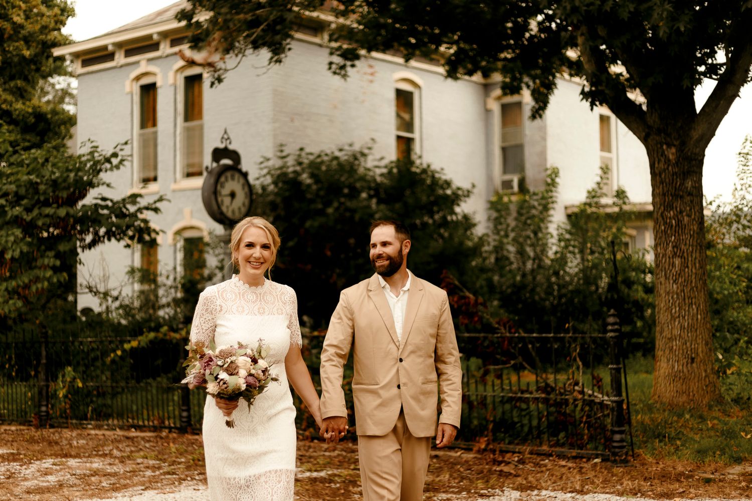 Bride and groom holding hands in front of a historic white mansion with a clock, bride holding a floral bouquet.