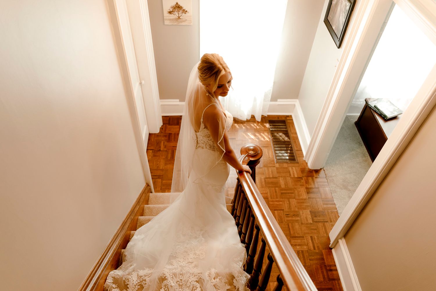 Bride in elegant white wedding gown and veil descending wooden staircase, viewed from above in a warmly lit home.