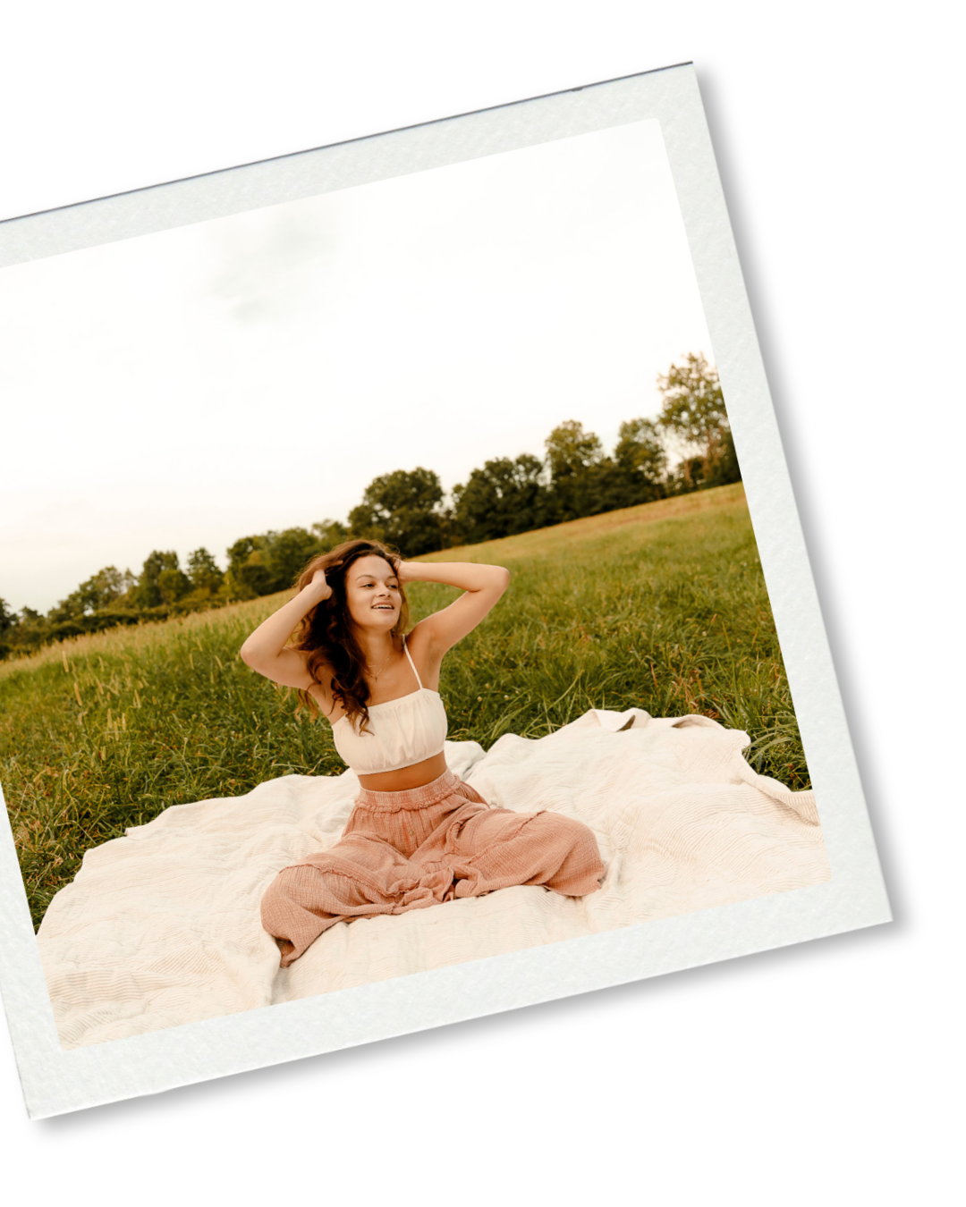 Woman in white crop top and pink pants sitting on blanket in green field, smiling with hands in hair.