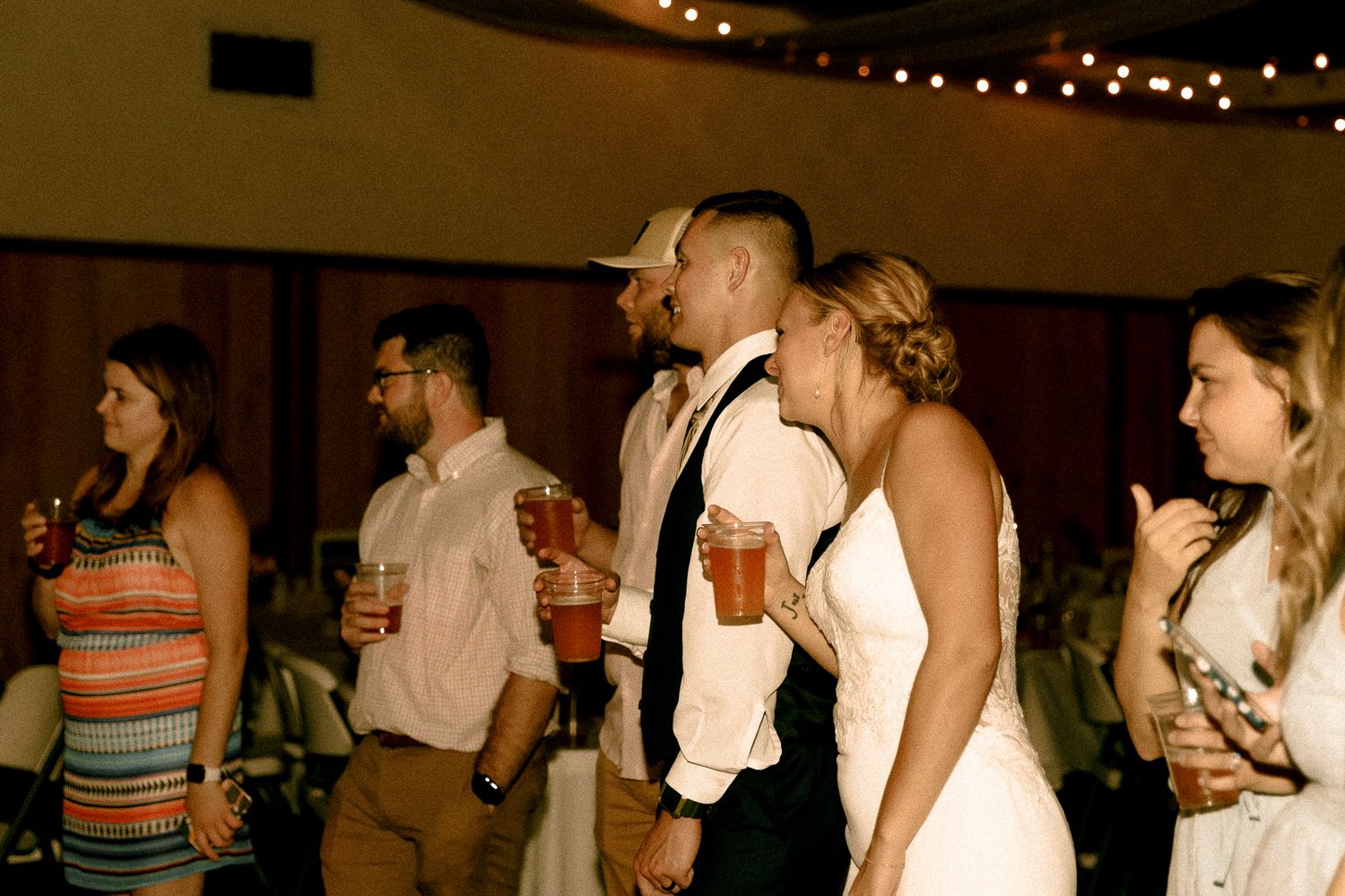 Wedding guests and bride in white gown socializing at indoor reception with string lights.