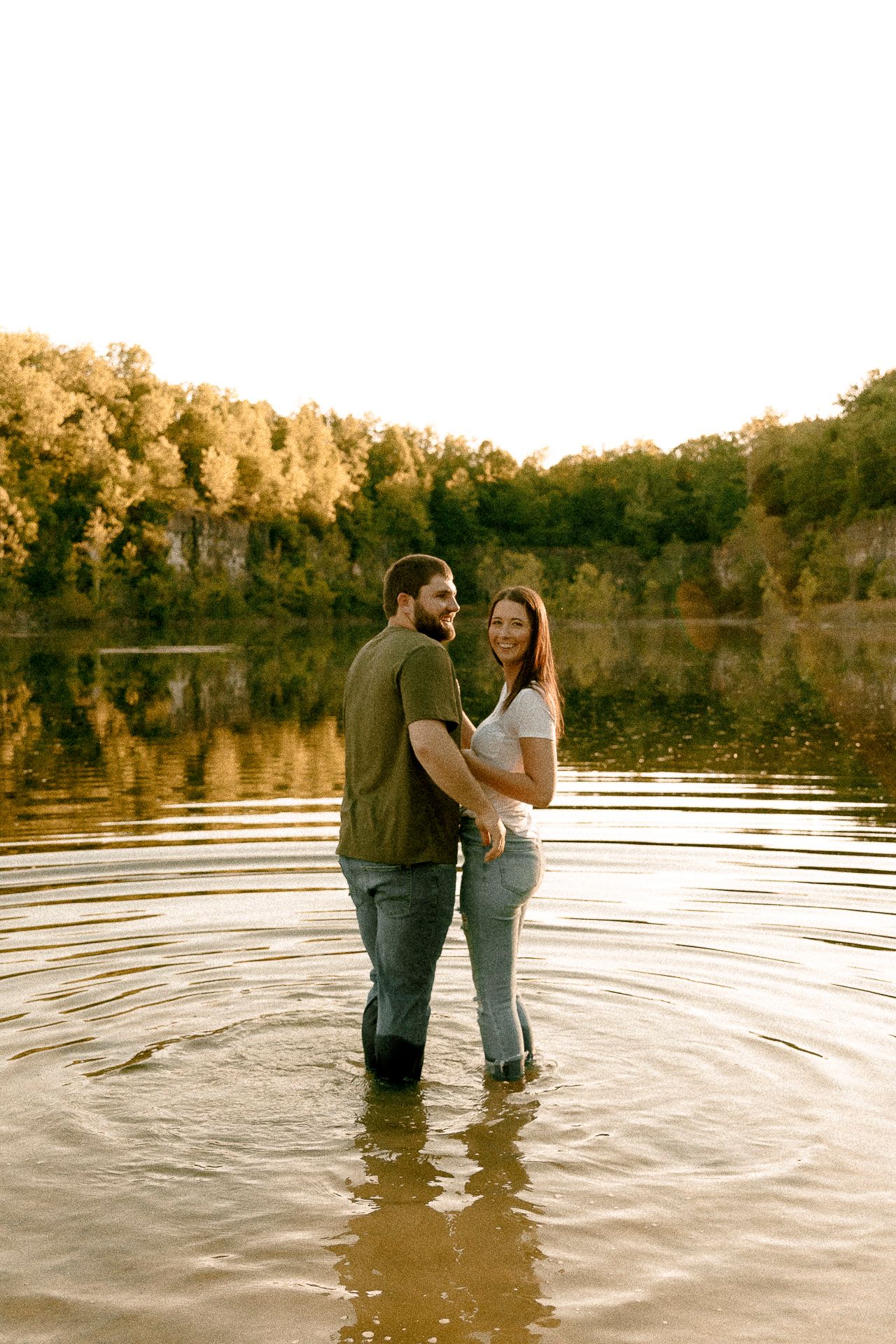 Couple standing in a lake during golden hour, embracing and smiling, surrounded by autumn trees and rocky cliffs.