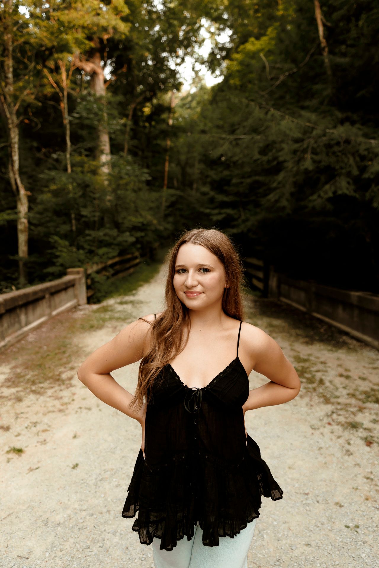 Young woman in black lace top poses on a sandy forest path surrounded by lush green trees.