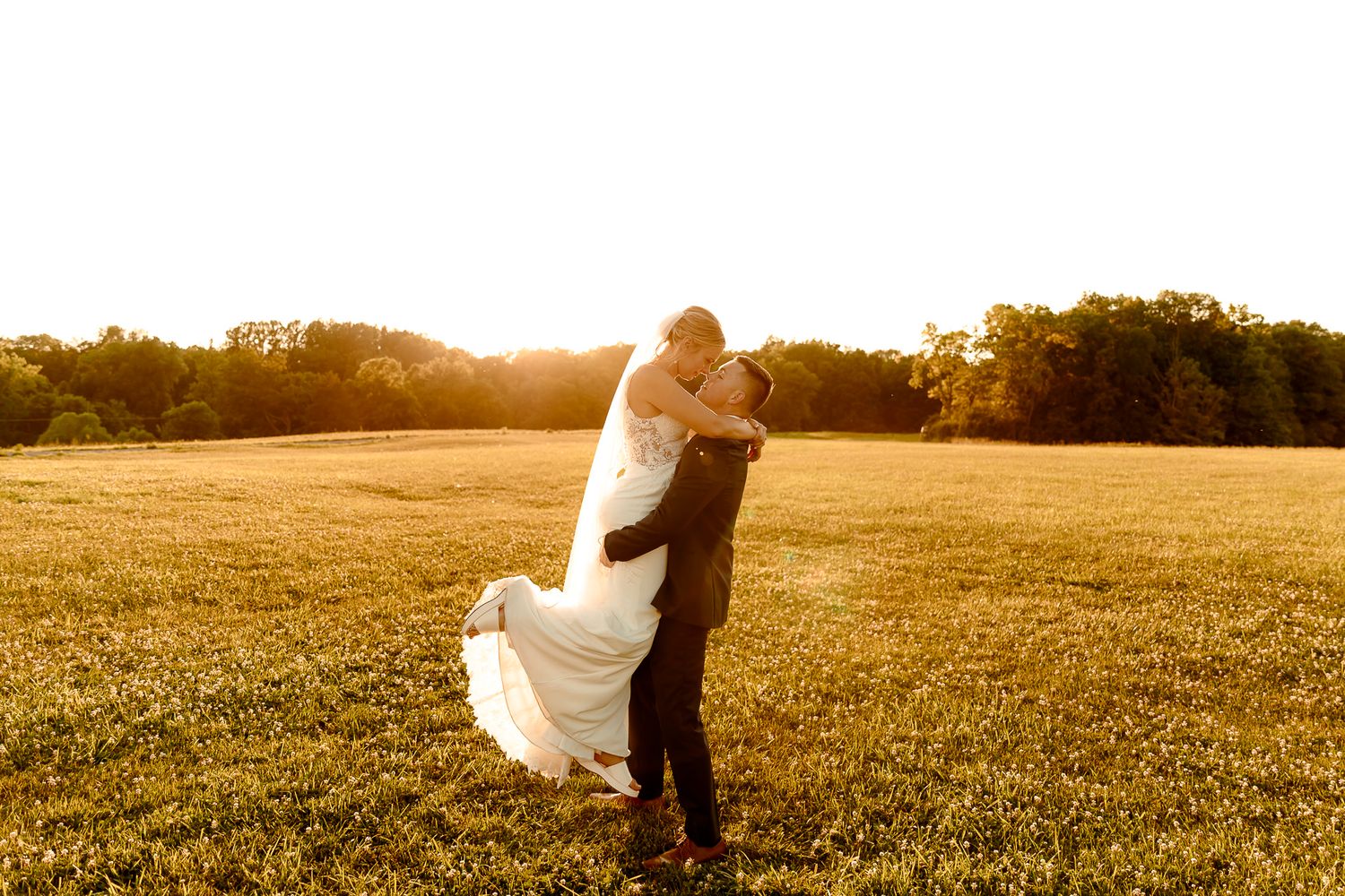 Groom lifting bride in white dress at golden sunset in a lush green field with trees in background.