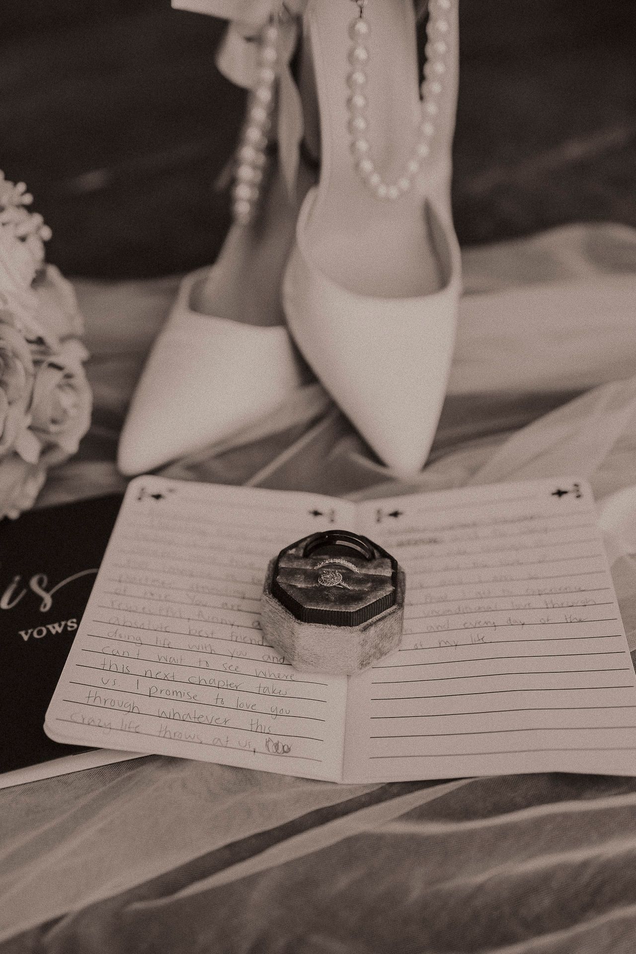 Black and white wedding flat lay with bridal heels, pearl necklace, ring box, and handwritten vows on open notepad.