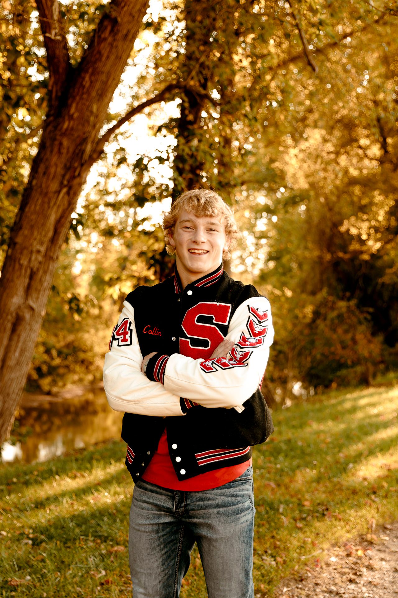 Smiling teenage boy in a varsity letterman jacket with arms crossed standing outdoors among autumn trees.