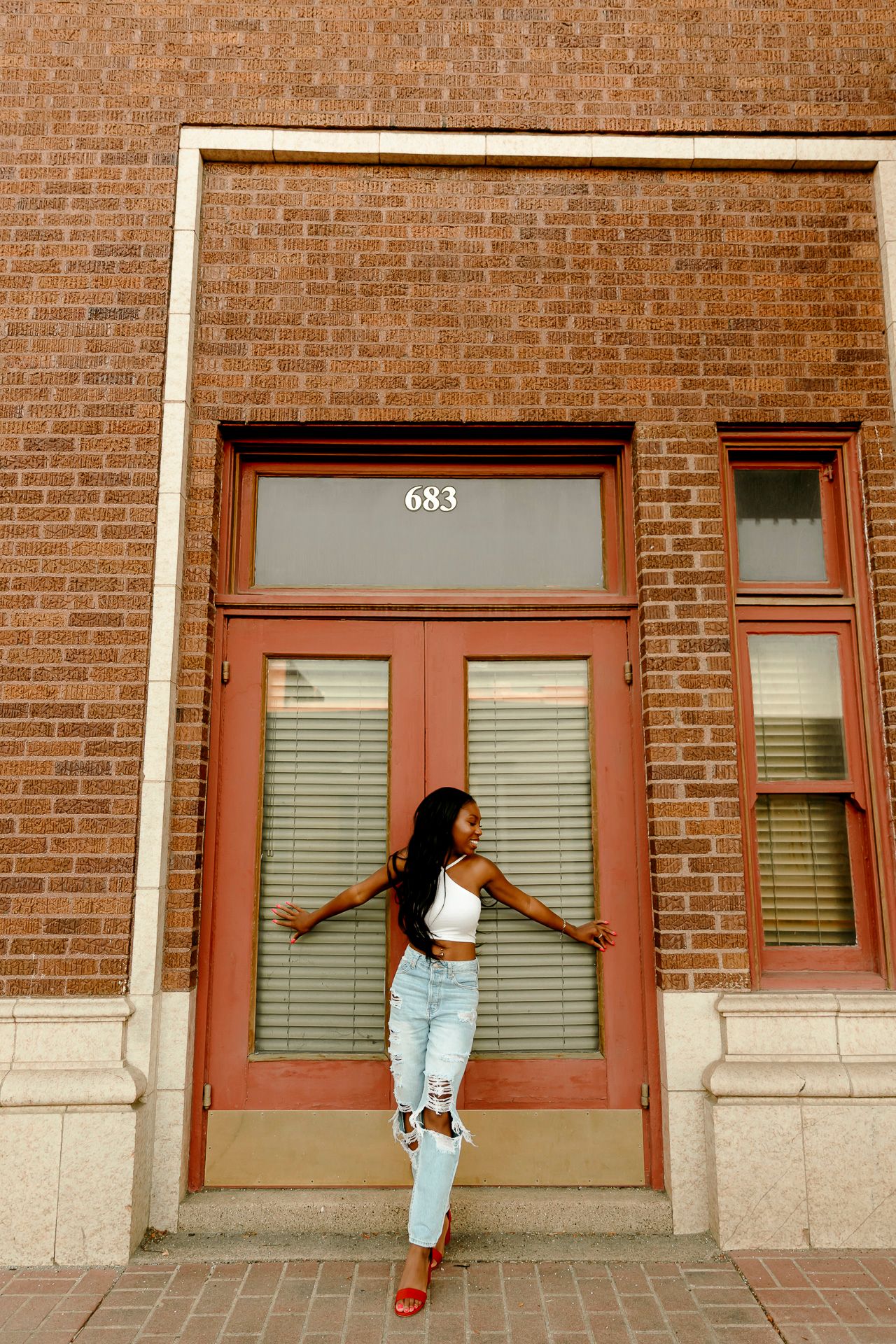 Young woman in white crop top and ripped jeans posing confidently in front of brick building entrance numbered 683.