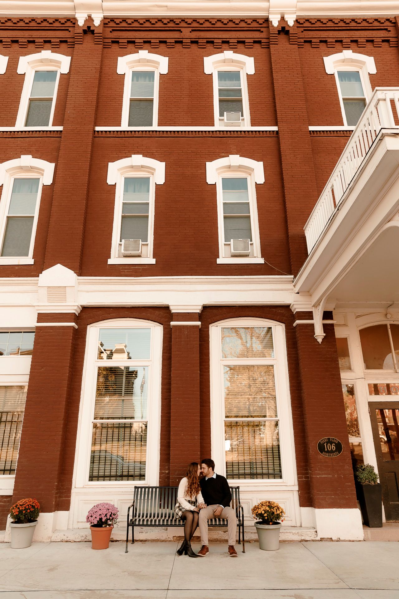 Couple sharing a romantic moment on a bench outside a historic red brick building with white trim and autumn flowers.