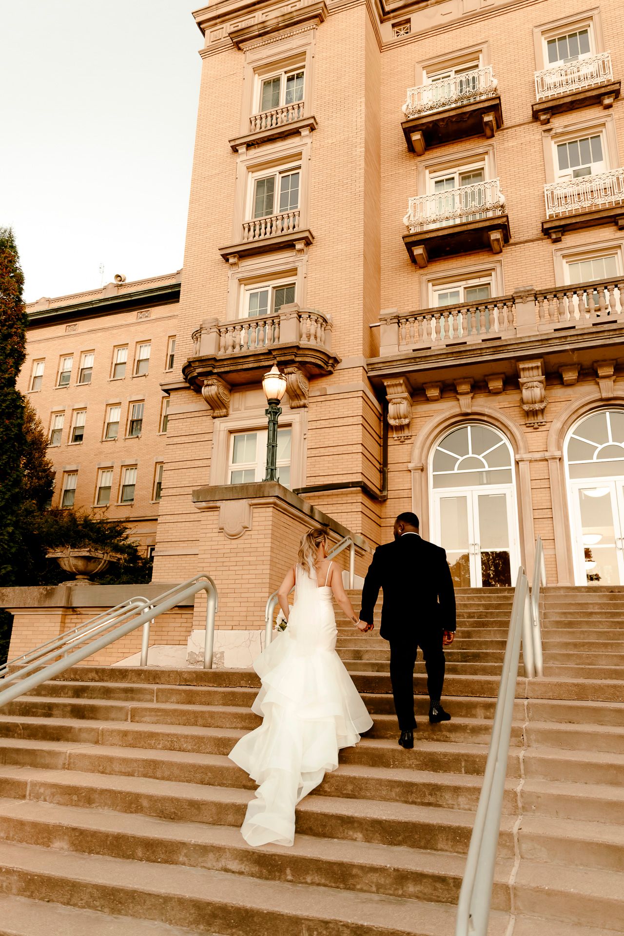 Bride and groom walk up grand stone steps toward elegant historic hotel building on their wedding day.
