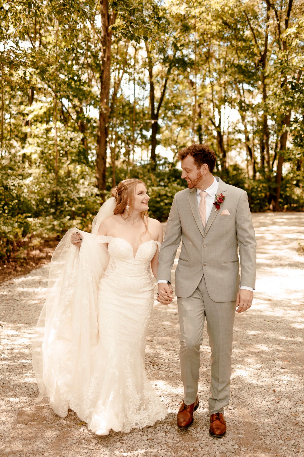 Bride and groom walk hand-in-hand on a tree-lined path, smiling at each other in wedding attire.