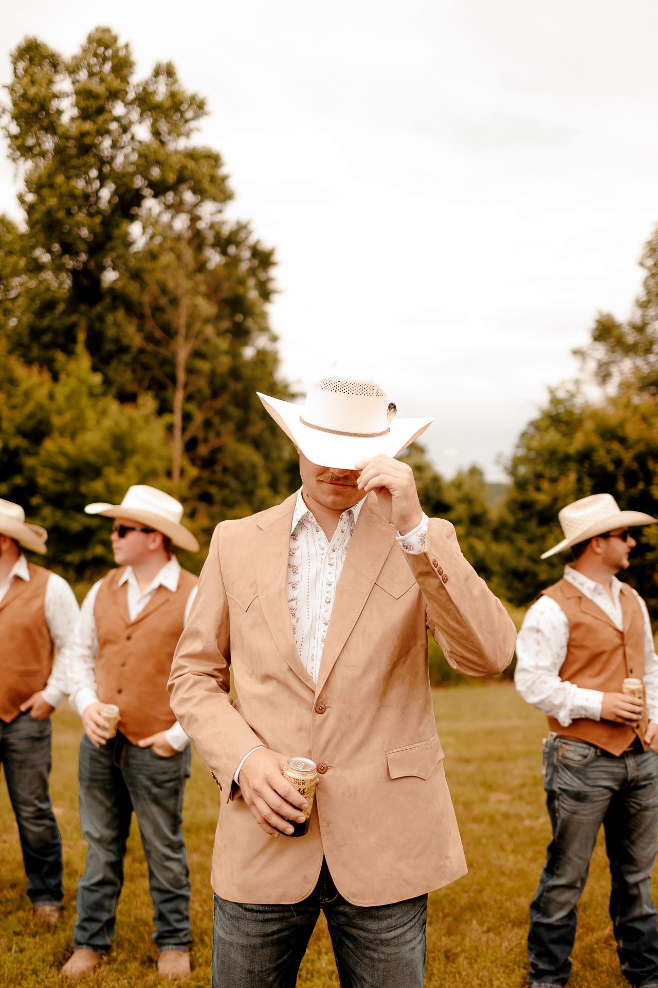 Groom in tan blazer and white cowboy hat with groomsmen in vests and cowboy hats at outdoor wedding.