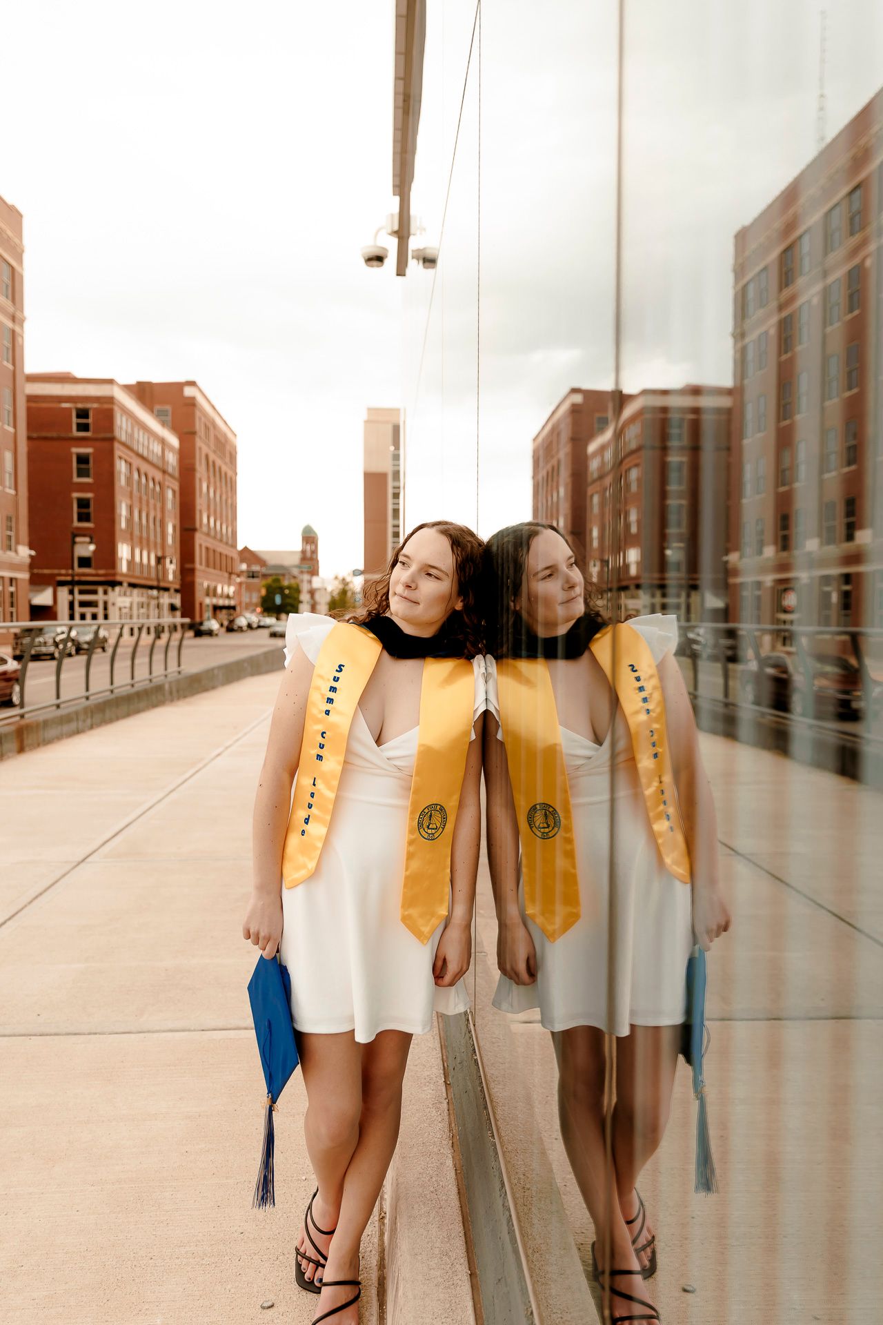 Graduate in white dress and gold sash leans against reflective glass wall, holding blue cap, with city buildings in background.