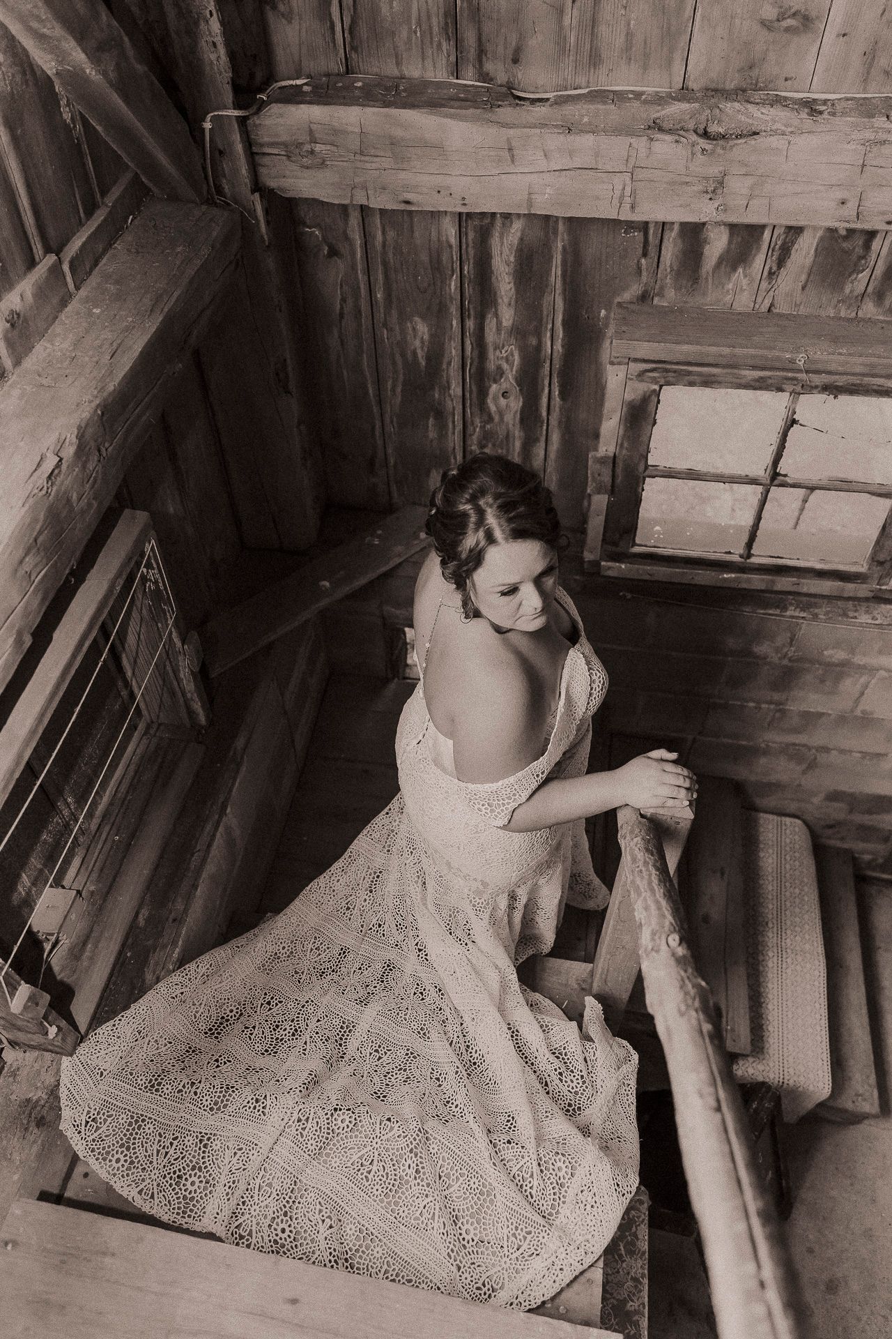 Bride in lace wedding dress descends rustic wooden staircase, sepia-toned photo capturing elegant vintage bridal style.