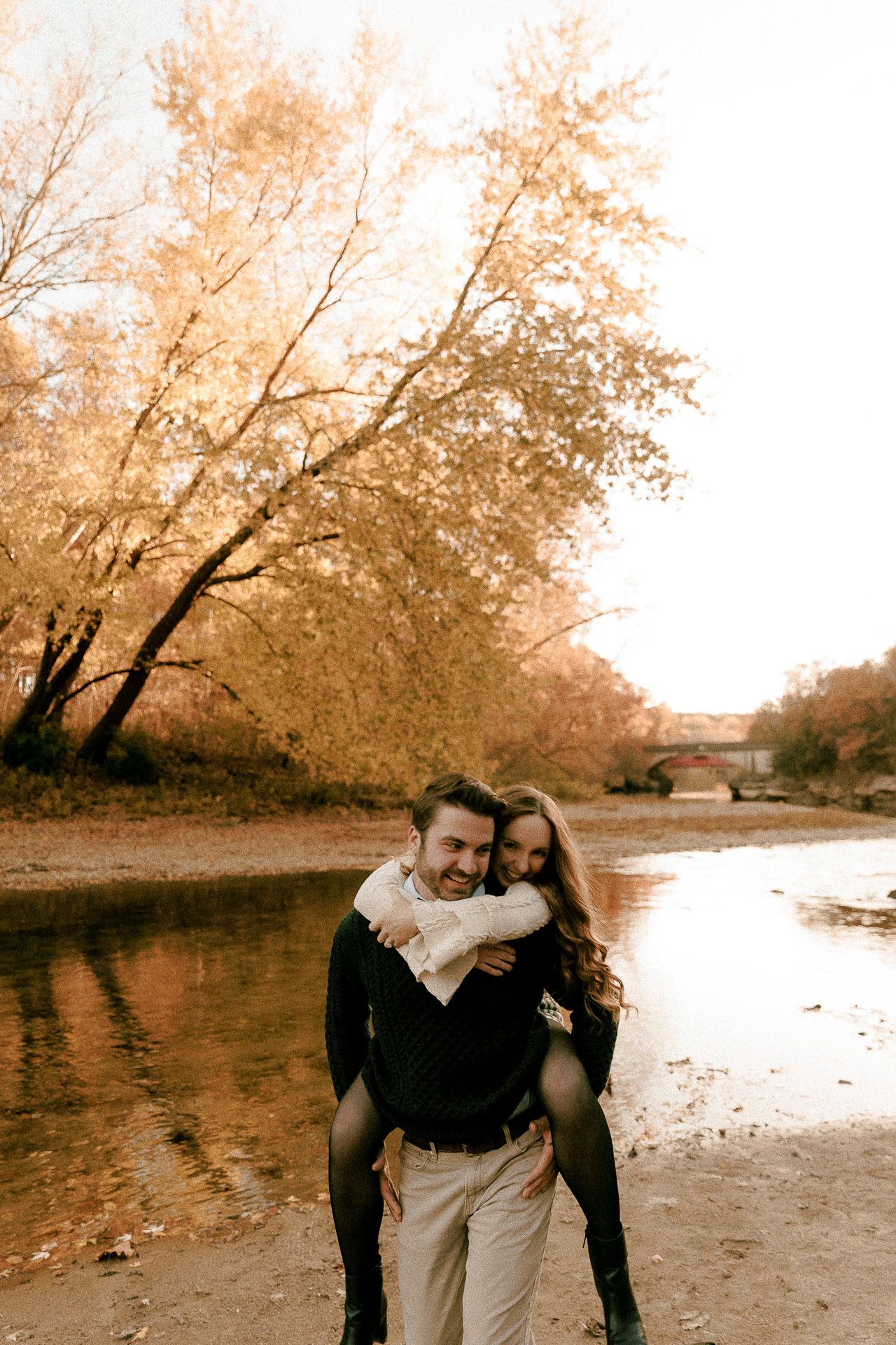 Couple sharing a piggyback ride by a river surrounded by golden autumn trees in a romantic outdoor setting.