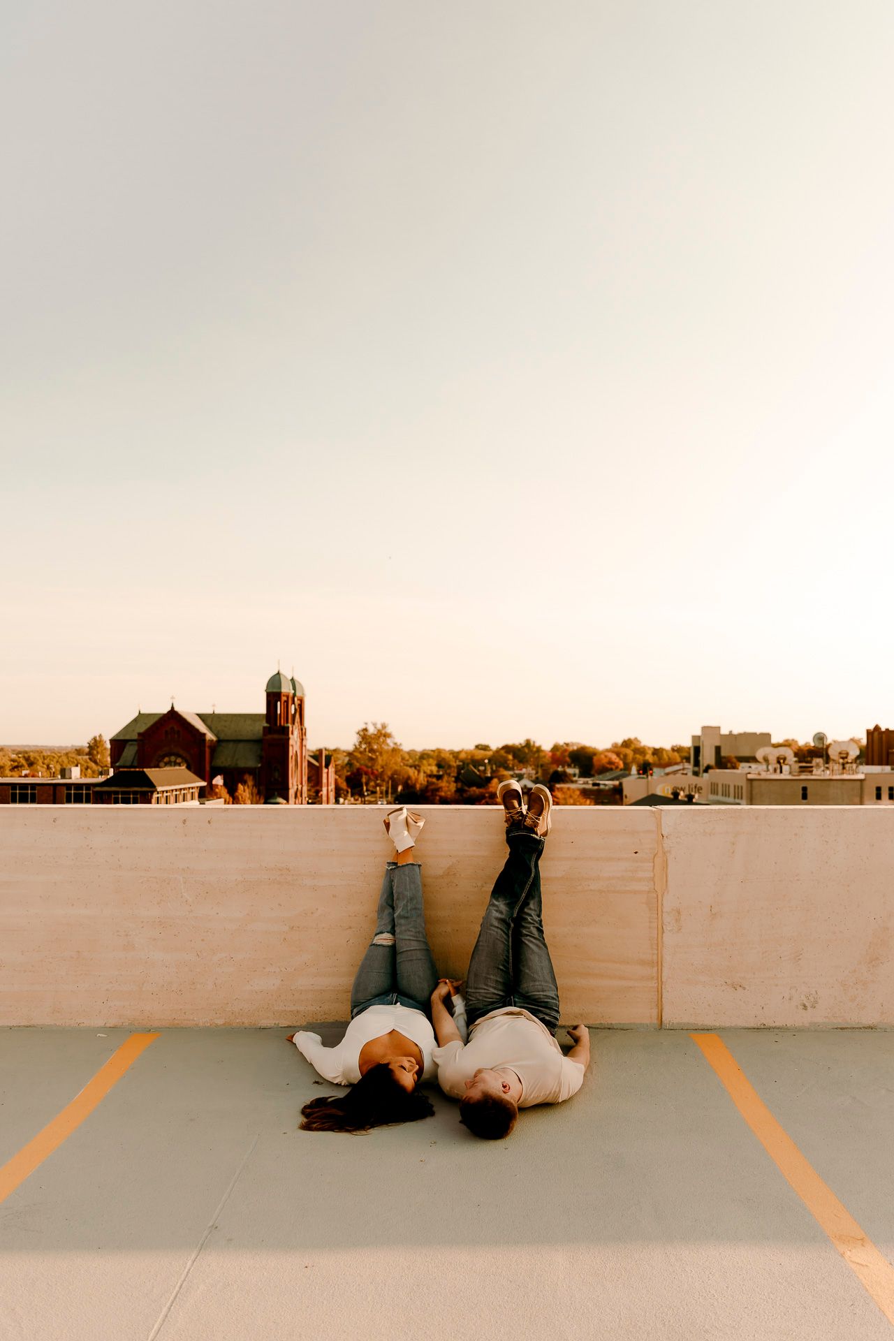 Two people lying on a rooftop parking deck with legs up against a wall, city skyline and church visible at sunset.