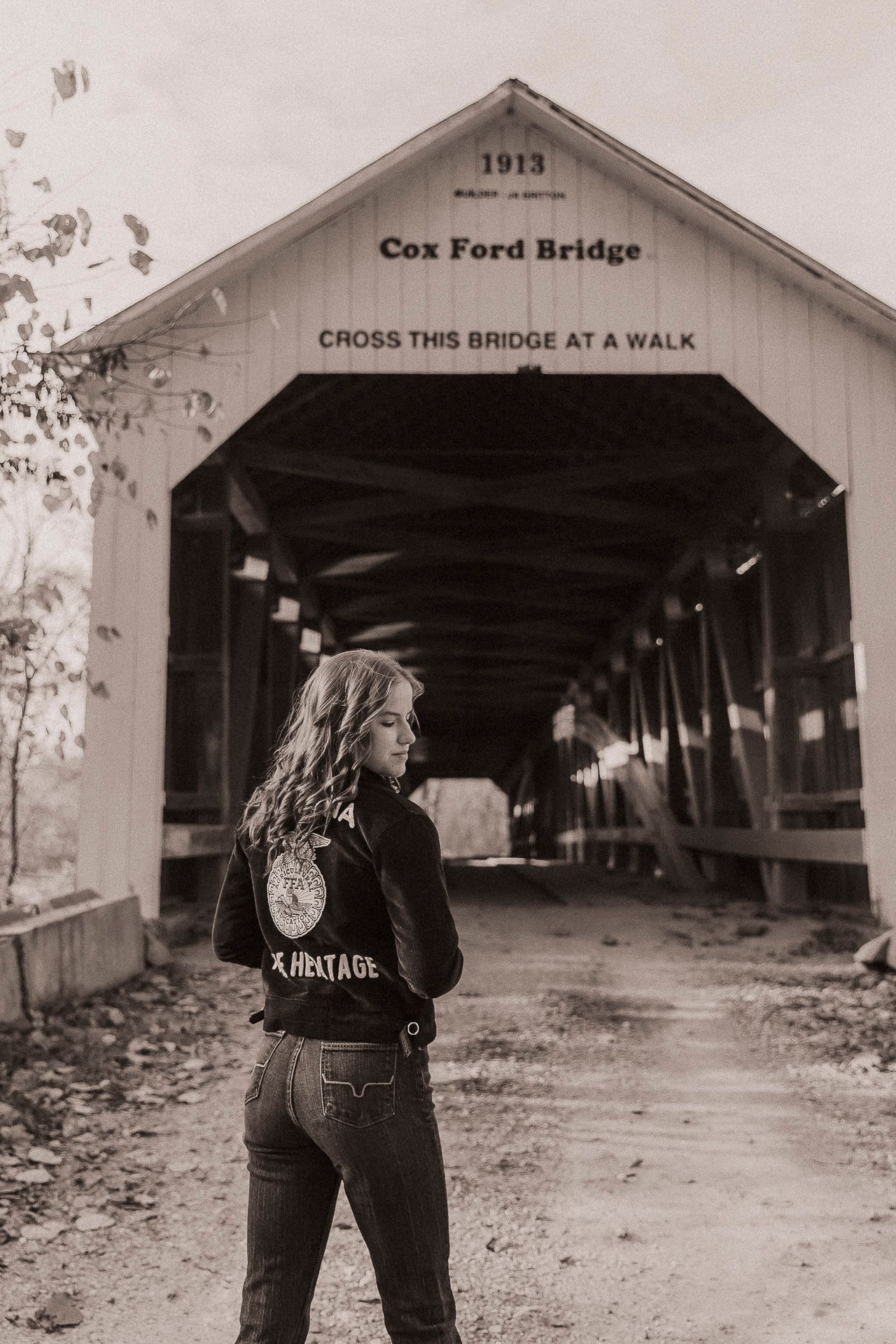Woman in heritage jacket stands before the historic 1913 Cox Ford covered bridge in a sepia-toned photo.