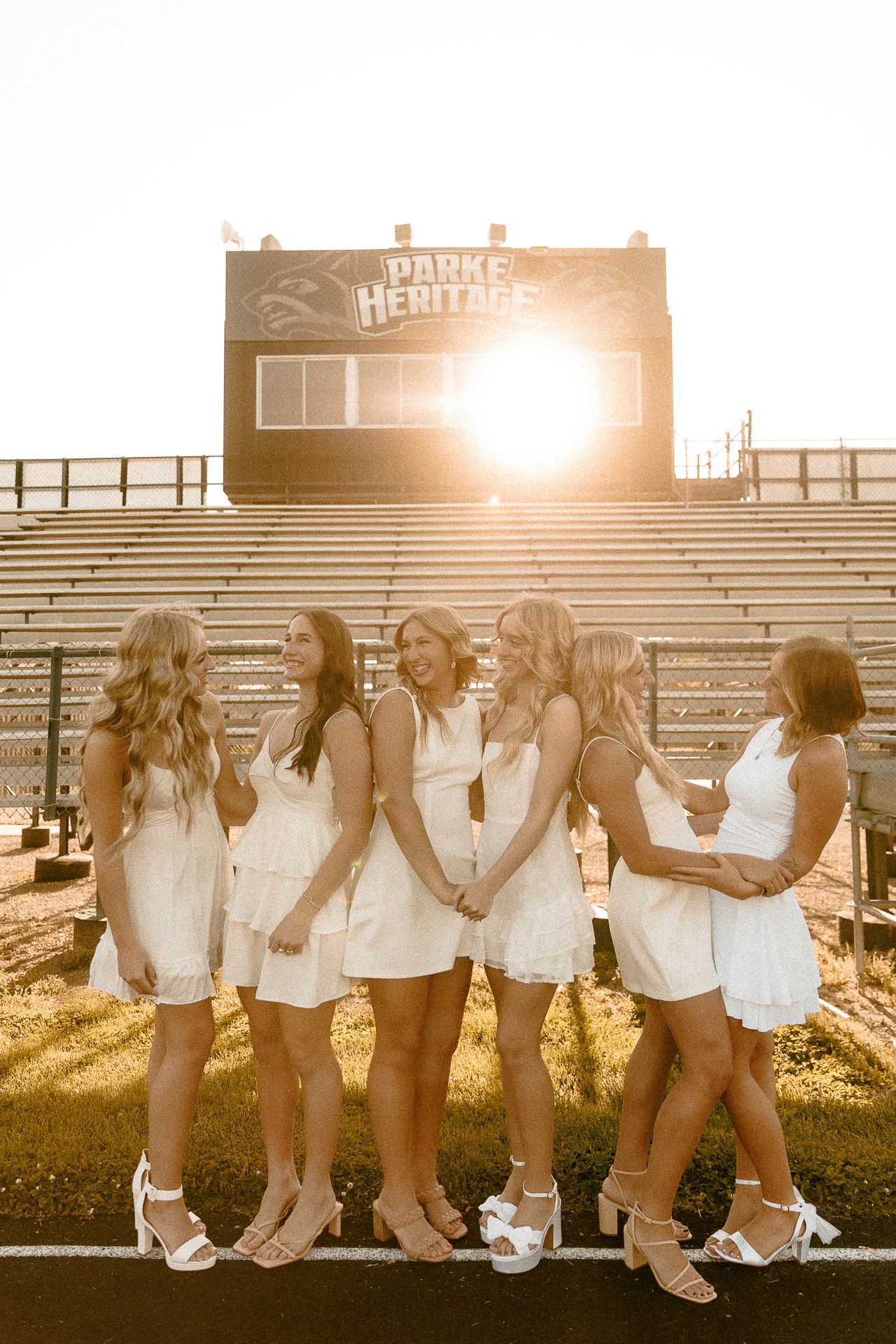 Six young women in white dresses pose together at golden hour in front of Parke Heritage stadium bleachers.