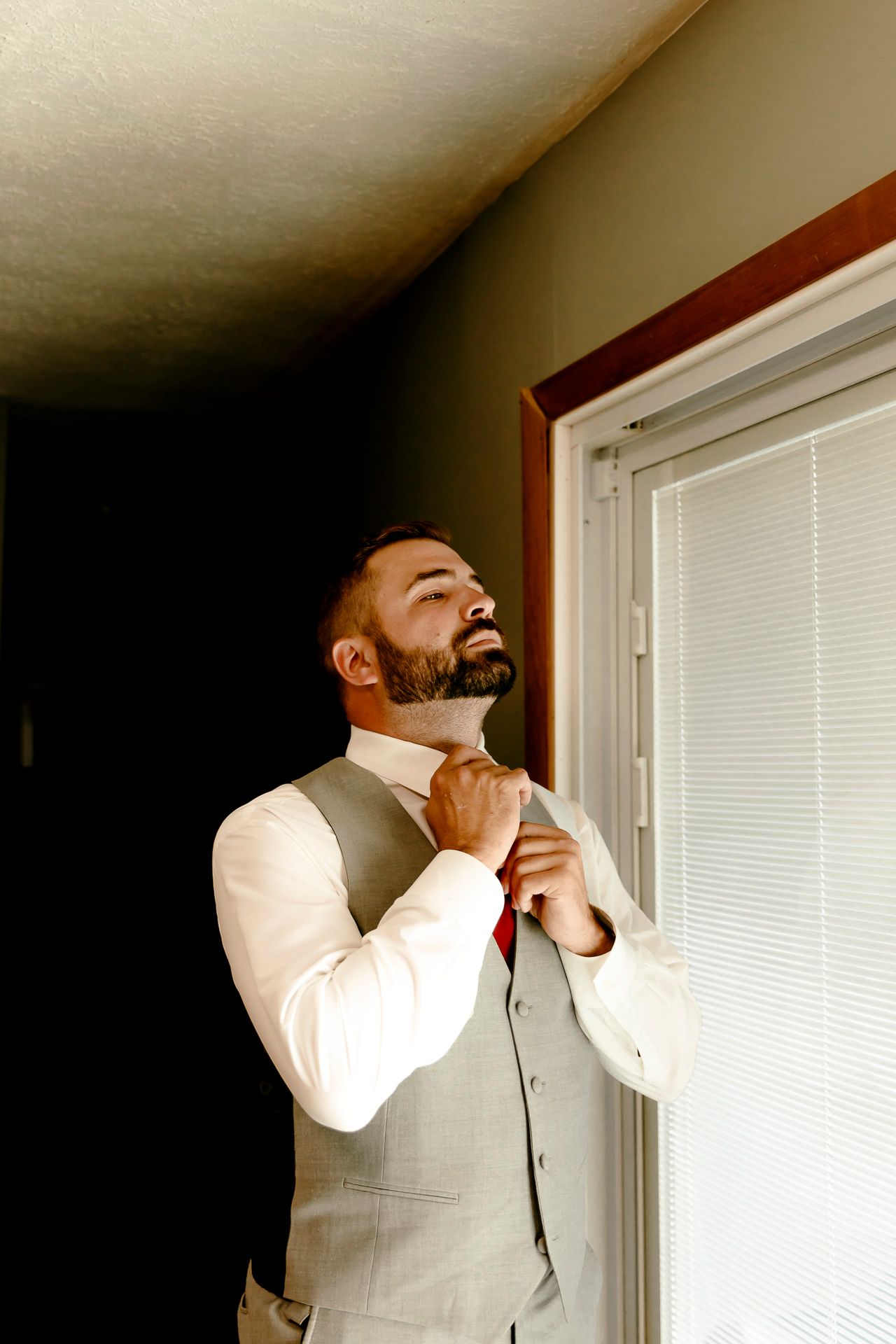 A bearded groom in a gray vest and red tie adjusts his collar while standing near a window with natural light.