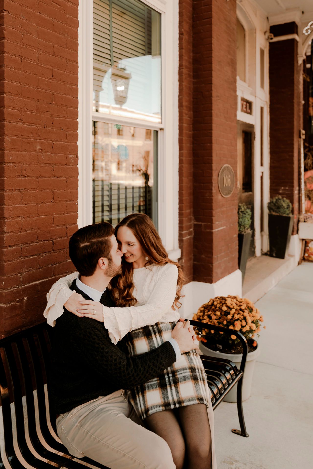 A couple sits closely on a bench outside a brick building, sharing a romantic moment with fall flowers nearby.