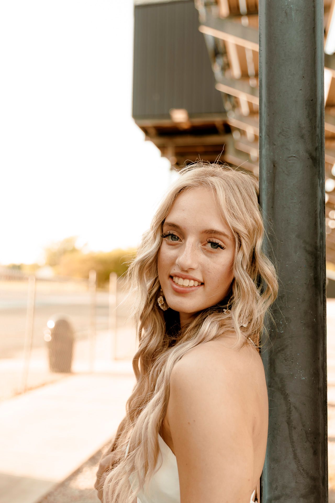 Smiling blonde teenage girl leaning against a metal pole outdoors in warm golden hour sunlight.
