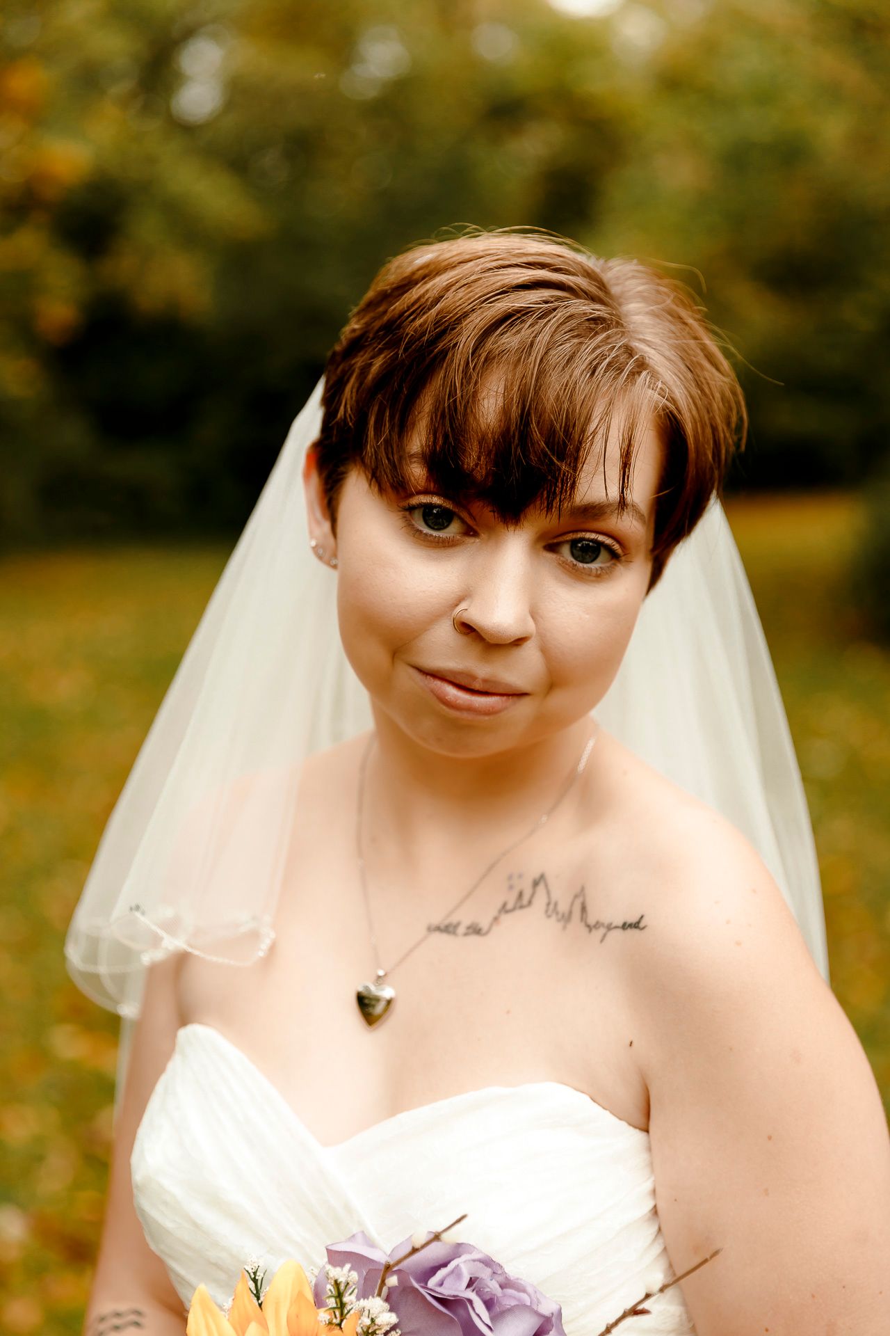 Bride with short brown hair wearing a white strapless gown, veil, and heart necklace holds a bouquet outdoors.