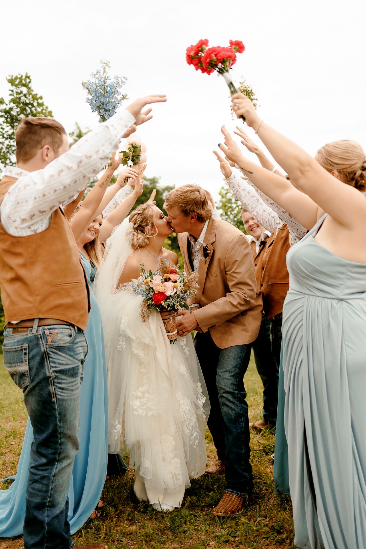 Bride and groom kiss under arch of raised hands with wedding party in blue and brown attire outdoors.