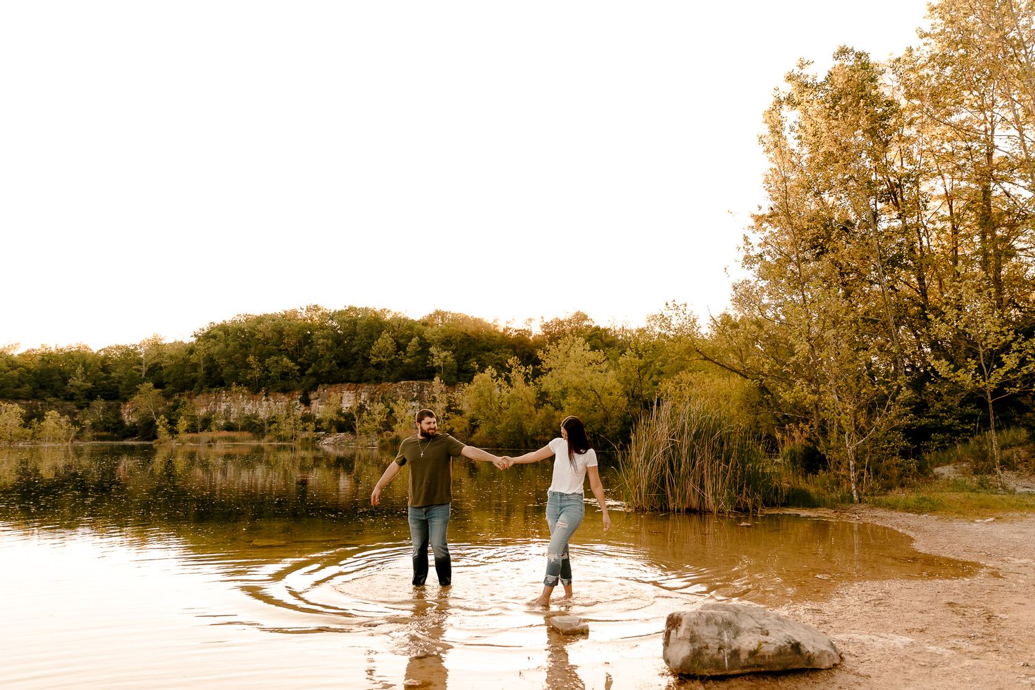 Couple holding hands wading in a shallow lake surrounded by autumn trees and rocky cliffs.