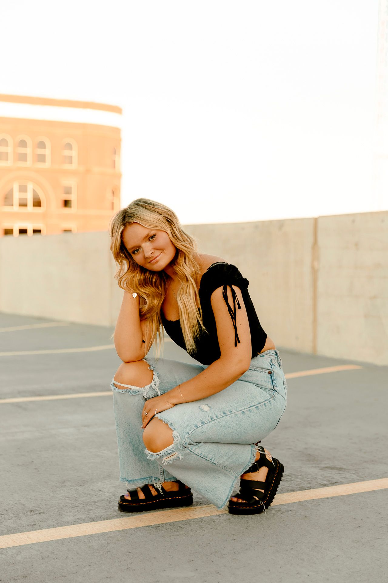 Blonde woman in black top and ripped jeans crouching on rooftop parking lot at golden hour.