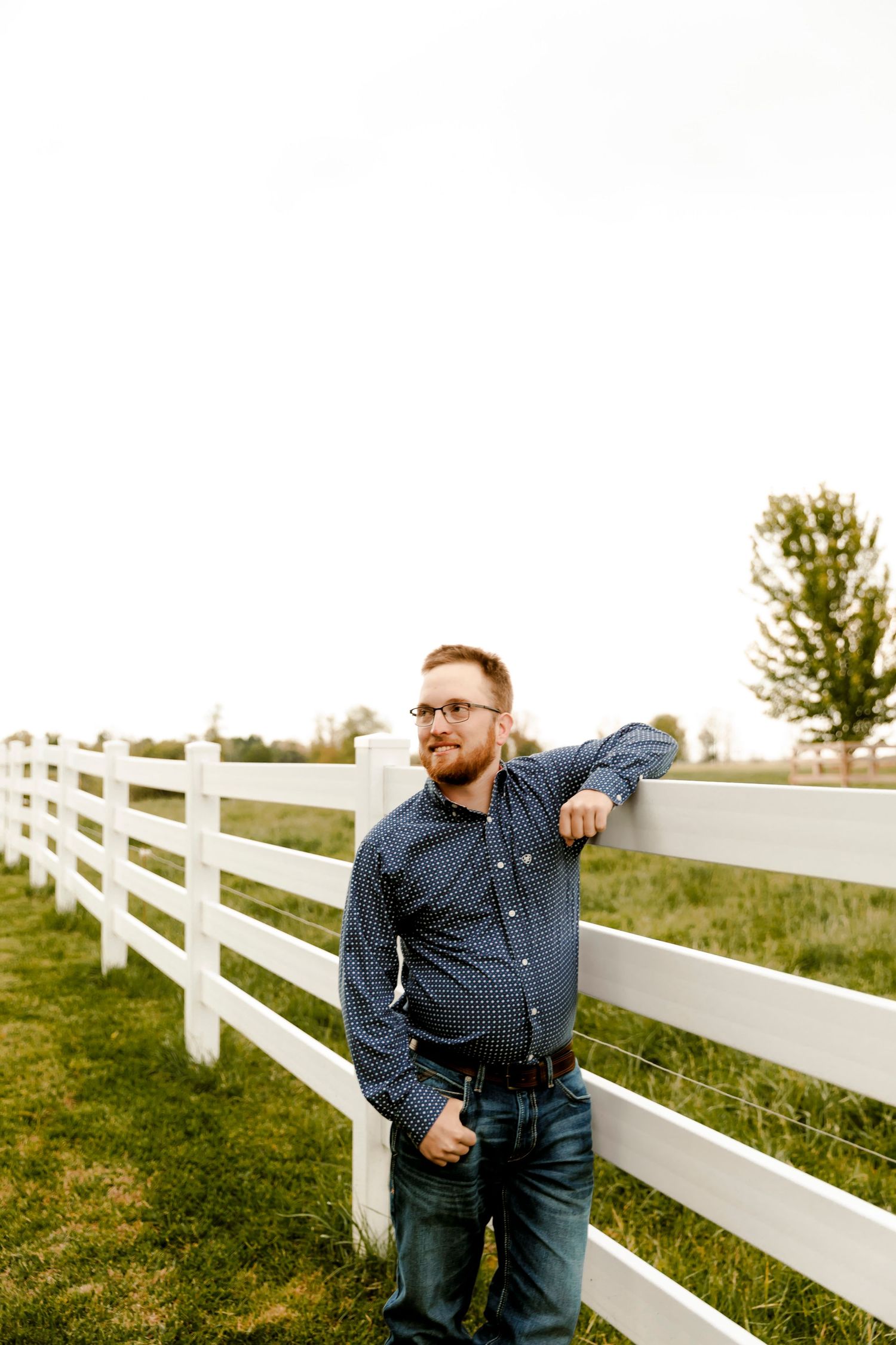 A bearded man in glasses and a blue shirt leans against a white farm fence in a green rural landscape.