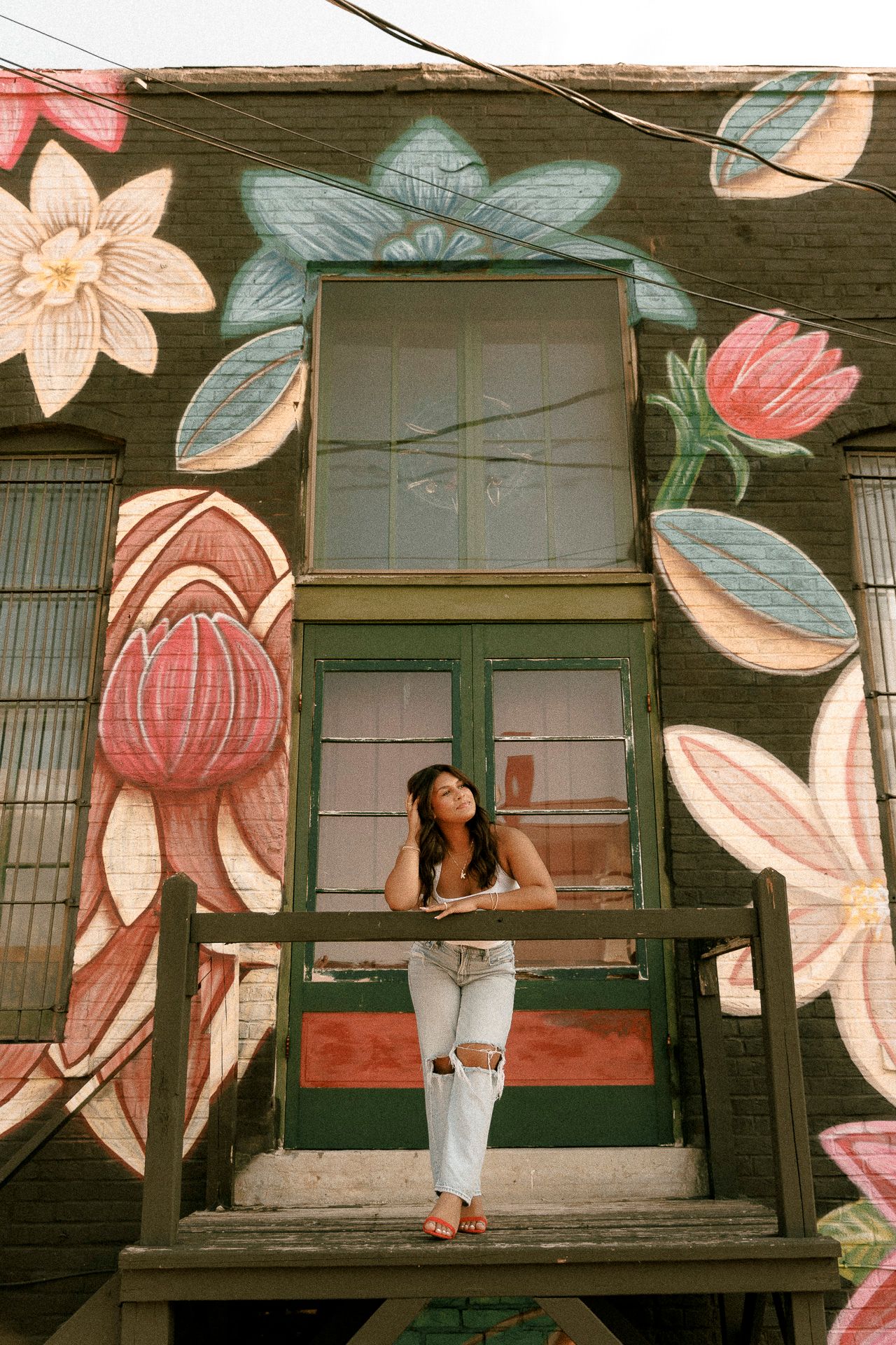 Woman in white jeans posing on a balcony against a colorful floral mural on a brick building.
