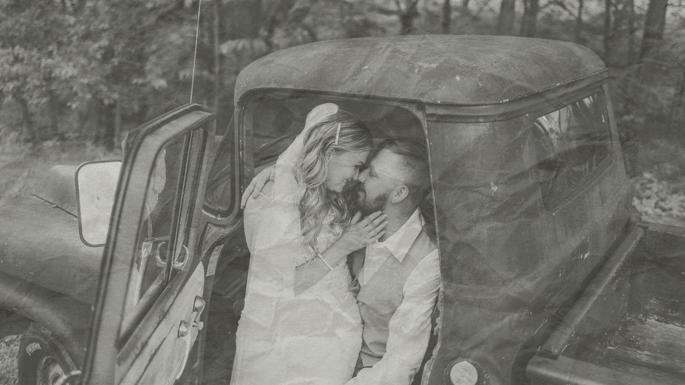 Black and white photo of a romantic couple sharing an intimate moment leaning against a vintage truck outdoors.