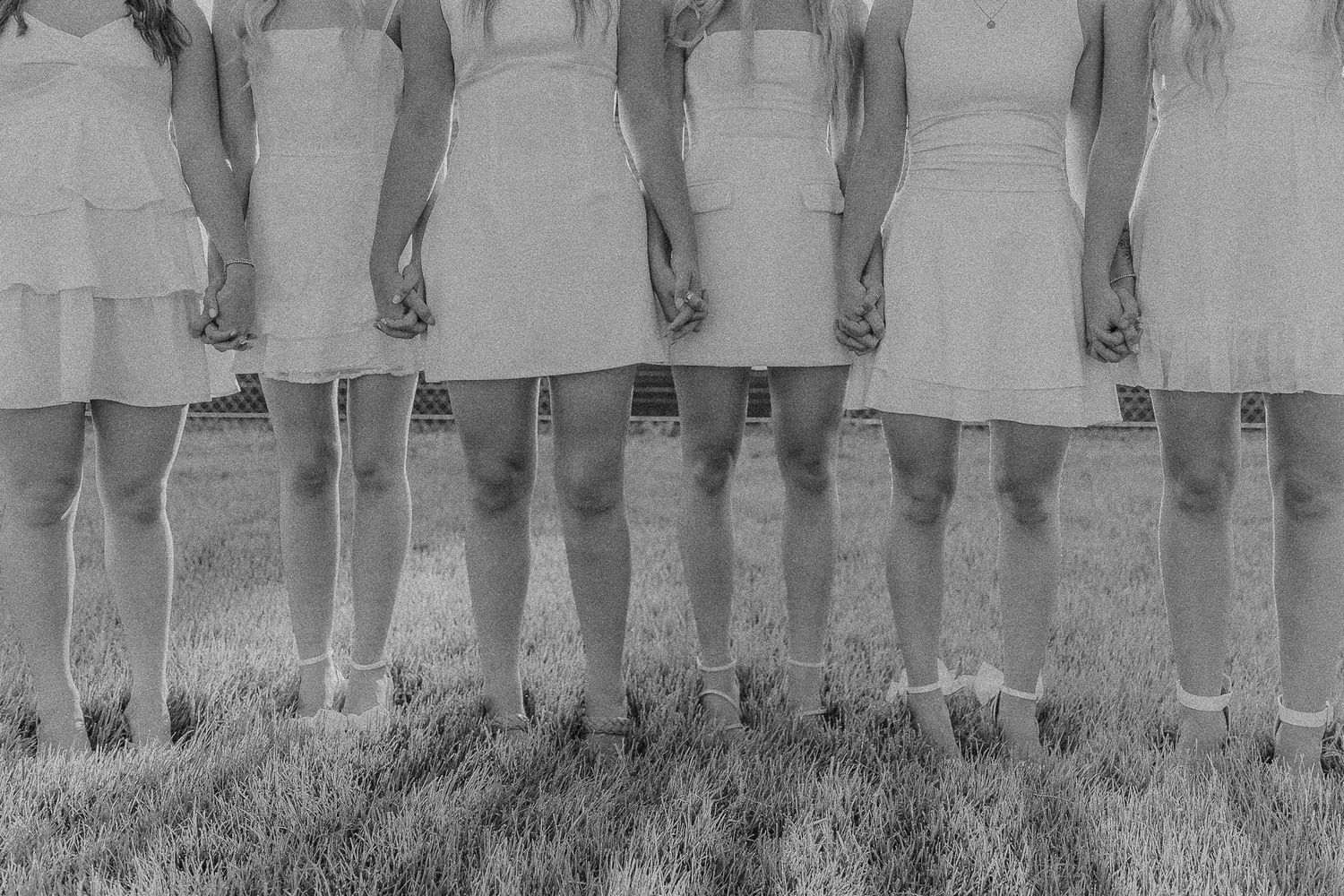 A group of bridesmaids in white dresses stand in a row outdoors, photographed from the waist down.