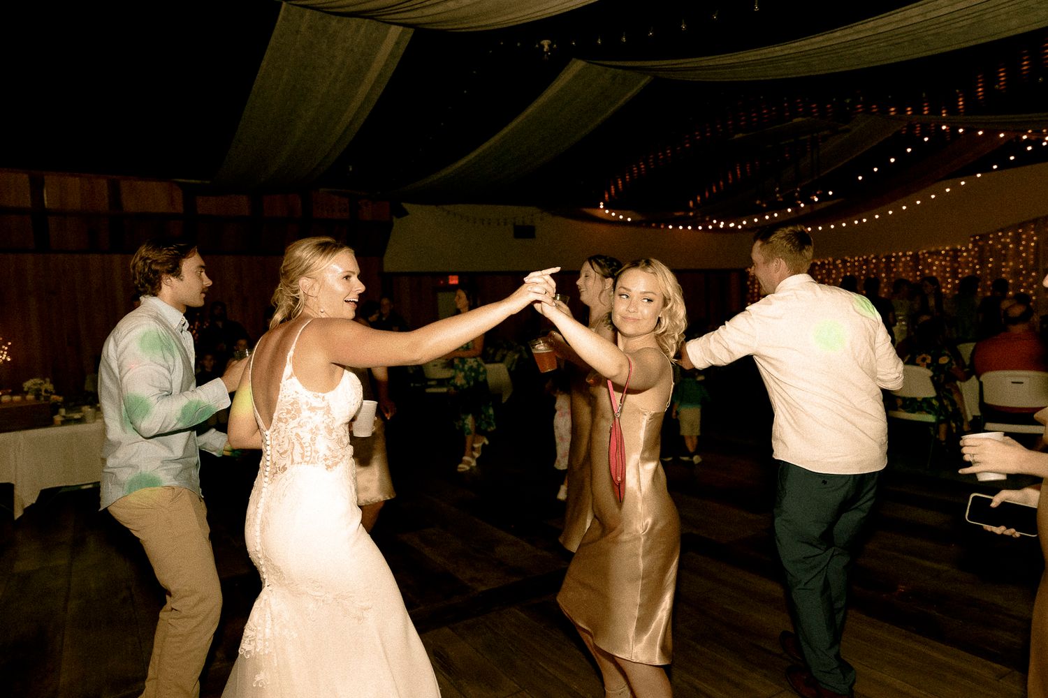Bride in white gown dancing with guests at elegant wedding reception with string lights.