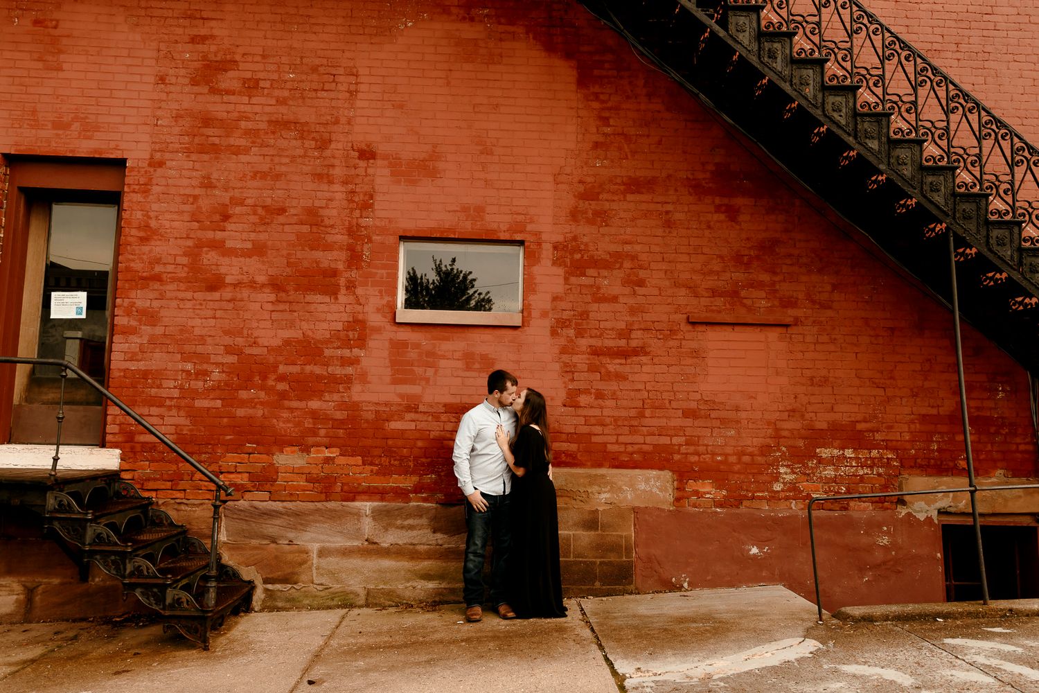 Couple sharing a romantic kiss against a rustic red brick wall with iron staircase in urban setting.