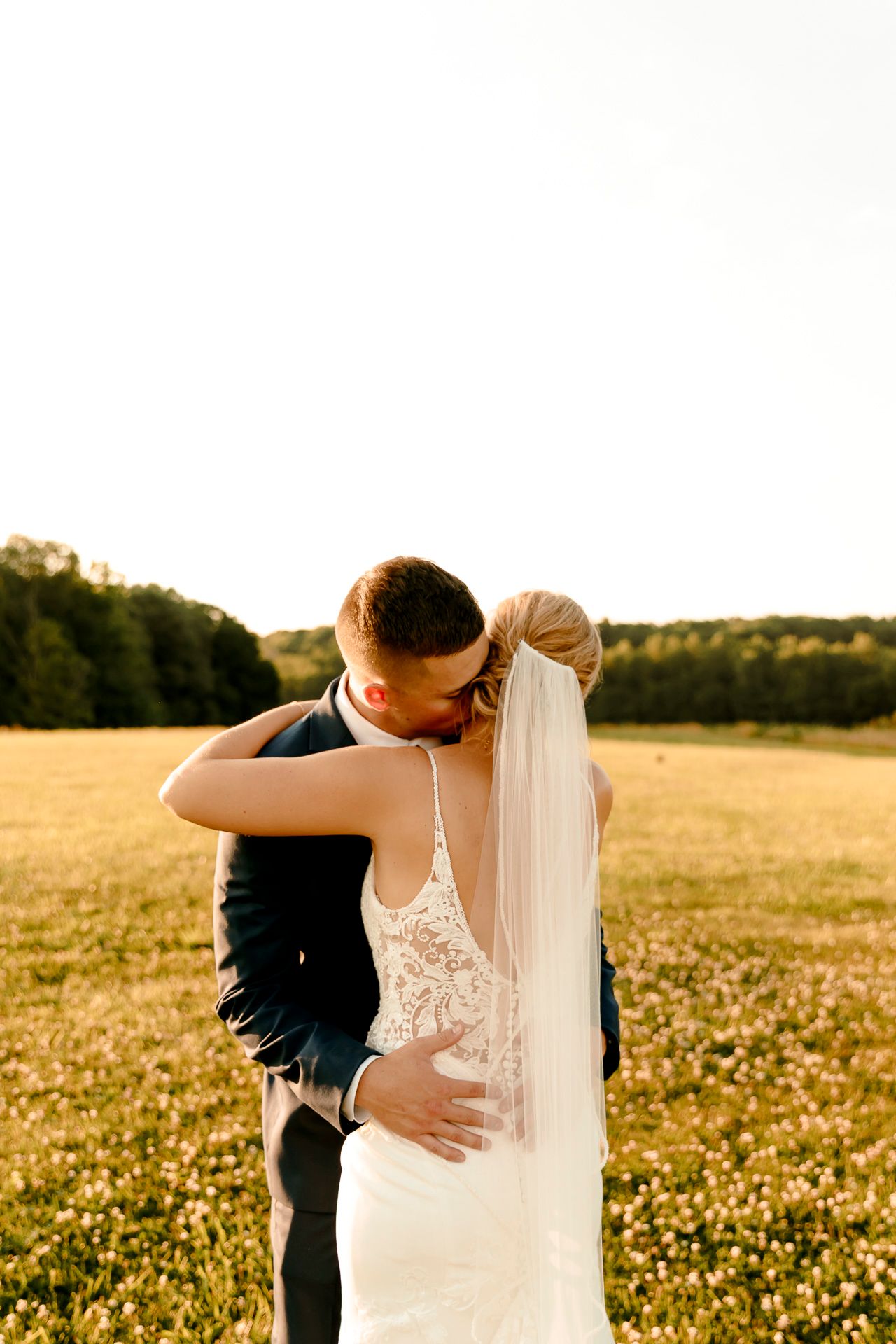Bride and groom sharing a romantic kiss in a golden sunlit field, bride wearing a lace gown and veil.