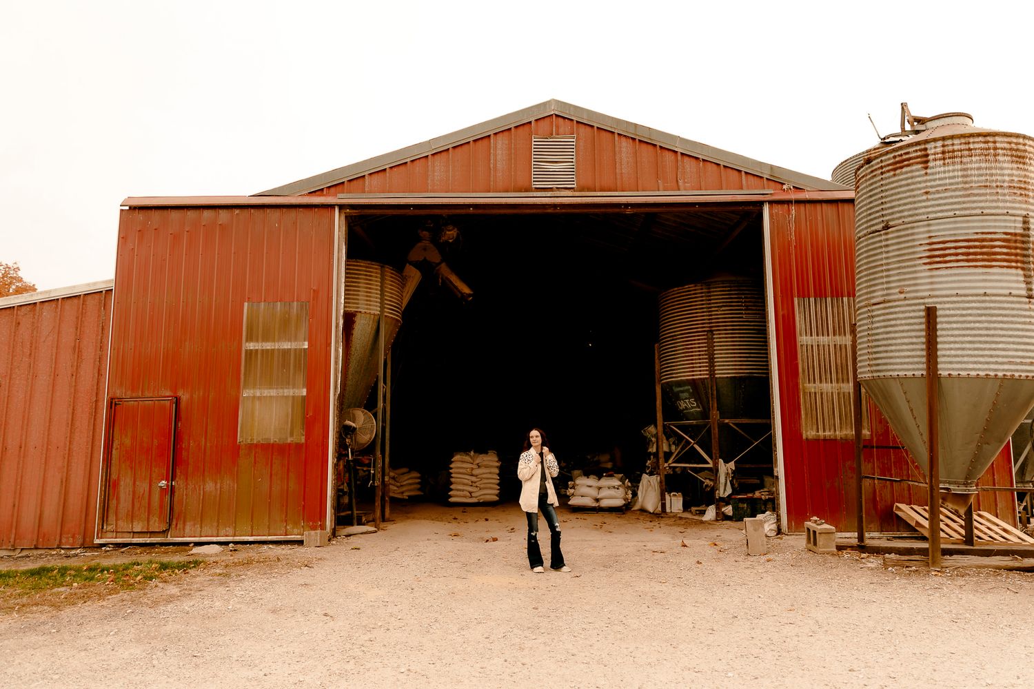 Person standing in front of a large red barn with open doors on a farm with grain silos.