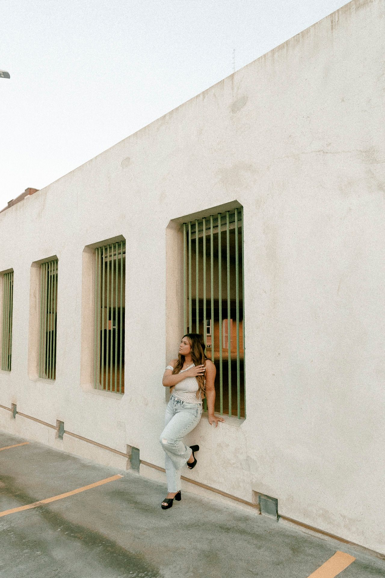 Woman in white top and jeans posing against a beige stucco wall with barred windows in a parking lot.