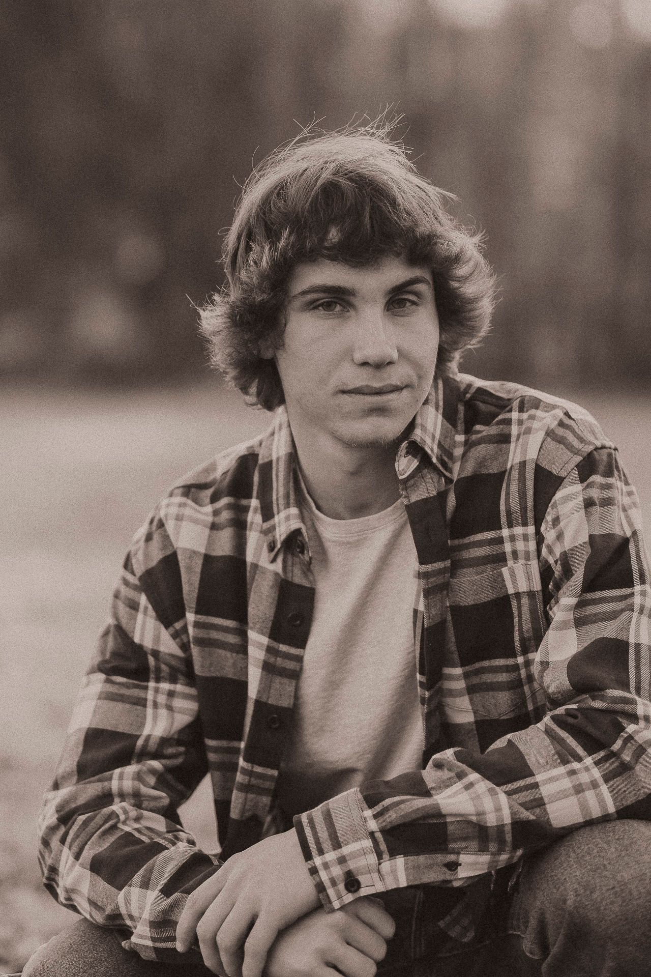 Black and white portrait of a young man in a plaid flannel shirt sitting outdoors with a natural background.