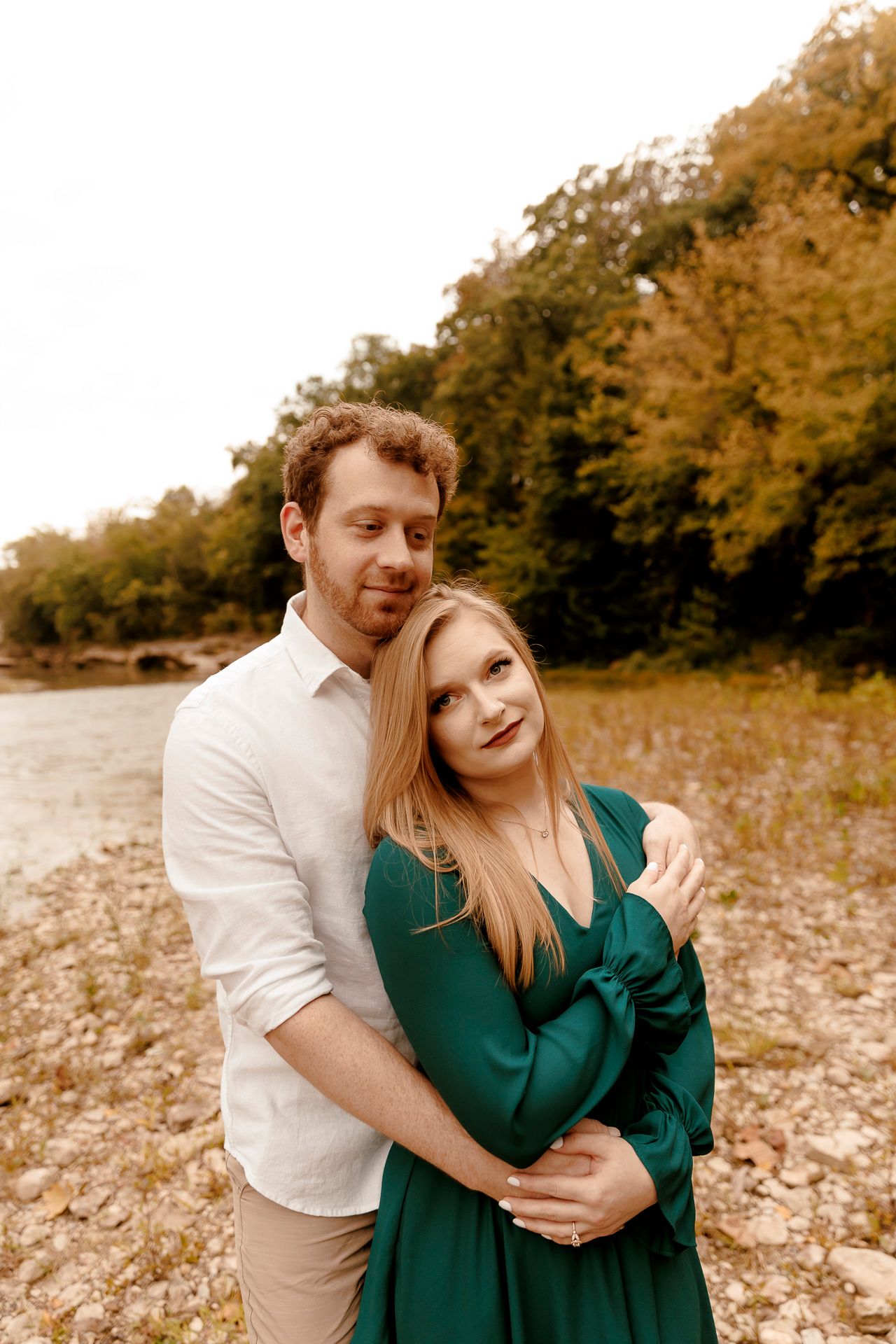 A couple embraces outdoors near a river, woman in green dress leaning on man in white shirt, with autumn trees behind.