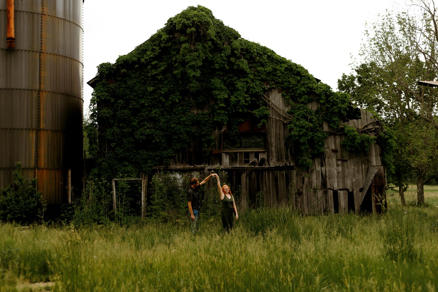 Two people walk through tall grass near an ivy-covered abandoned barn and grain silo on a rural farm.