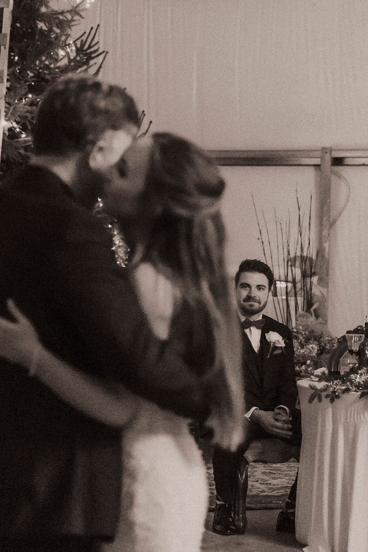 Black and white wedding photo of couple kissing while groomsman watches in background at elegant reception.