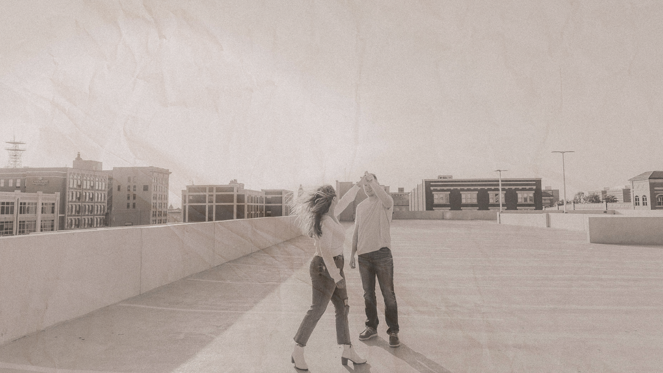 A couple dancing together on a rooftop parking structure with urban buildings in the background.