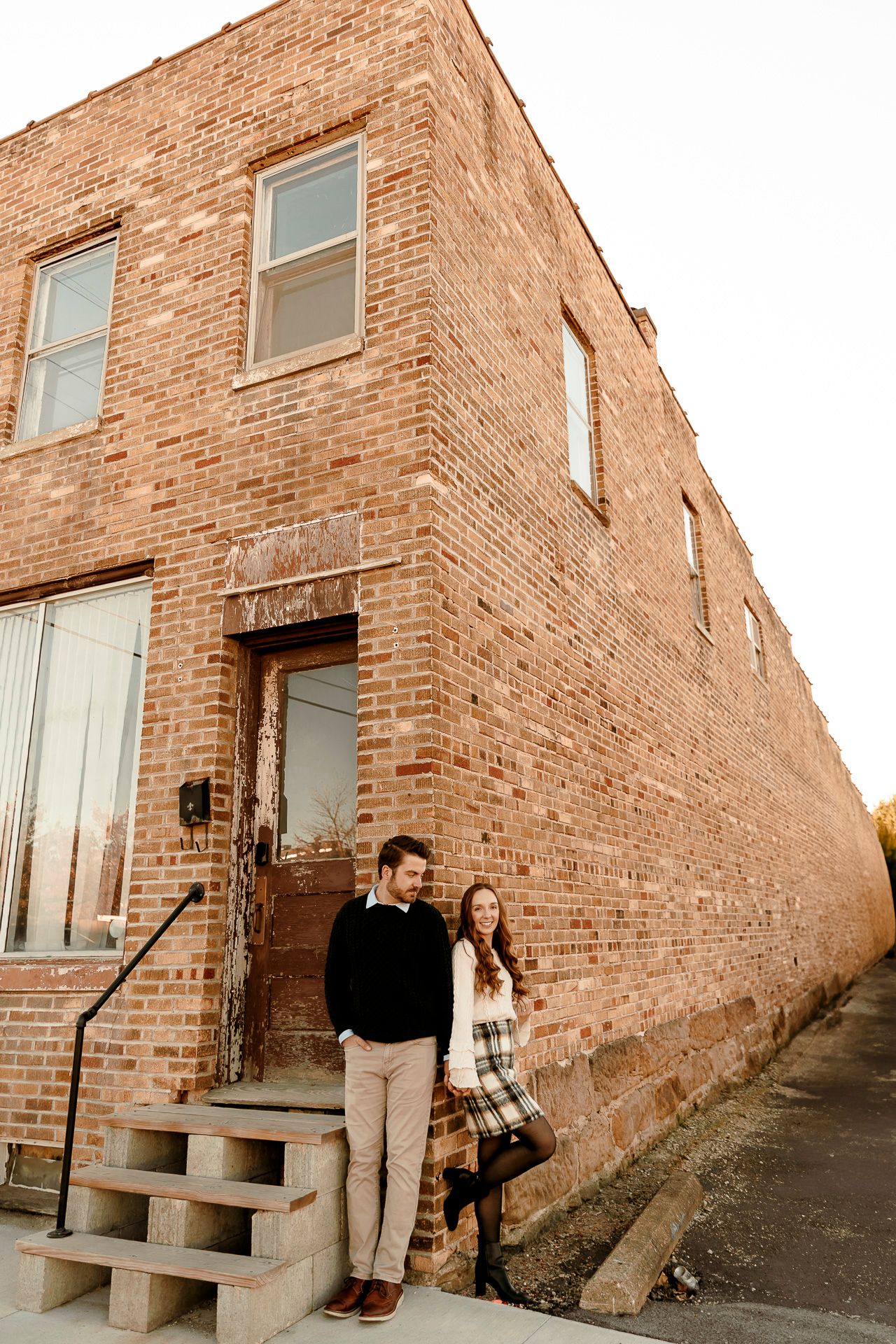 Couple posing together outside a rustic brick building with concrete steps during golden hour.
