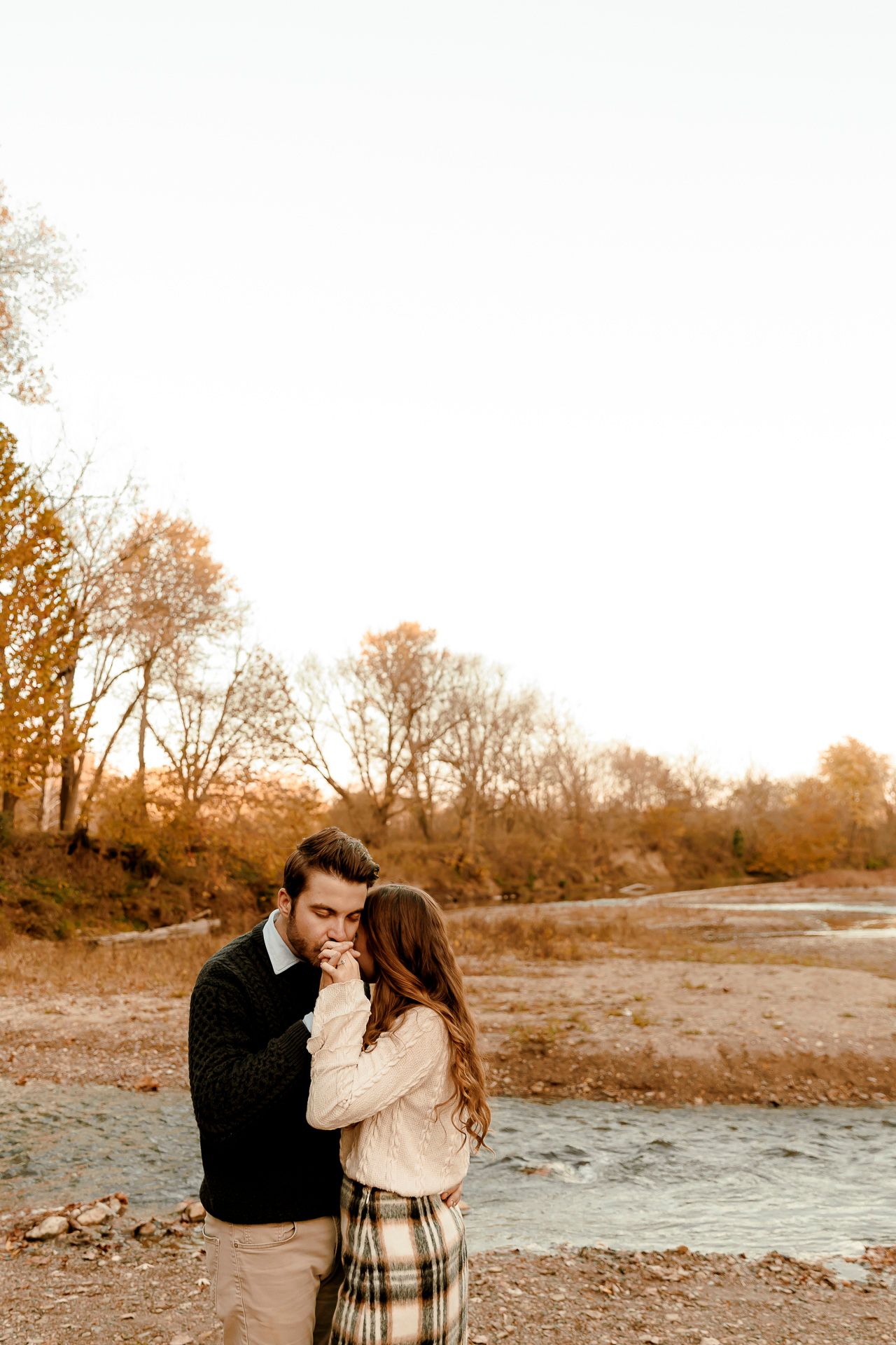 Couple sharing a romantic moment by a river in autumn, surrounded by golden trees and warm fall colors.