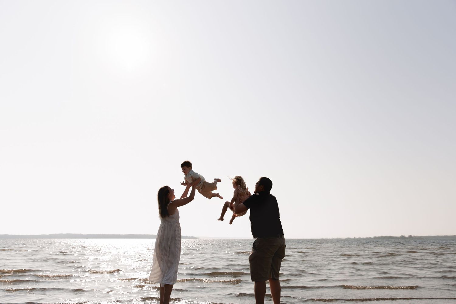 Family joyfully tossing two babies in the air on a sunny beach with calm ocean waters in the background.
