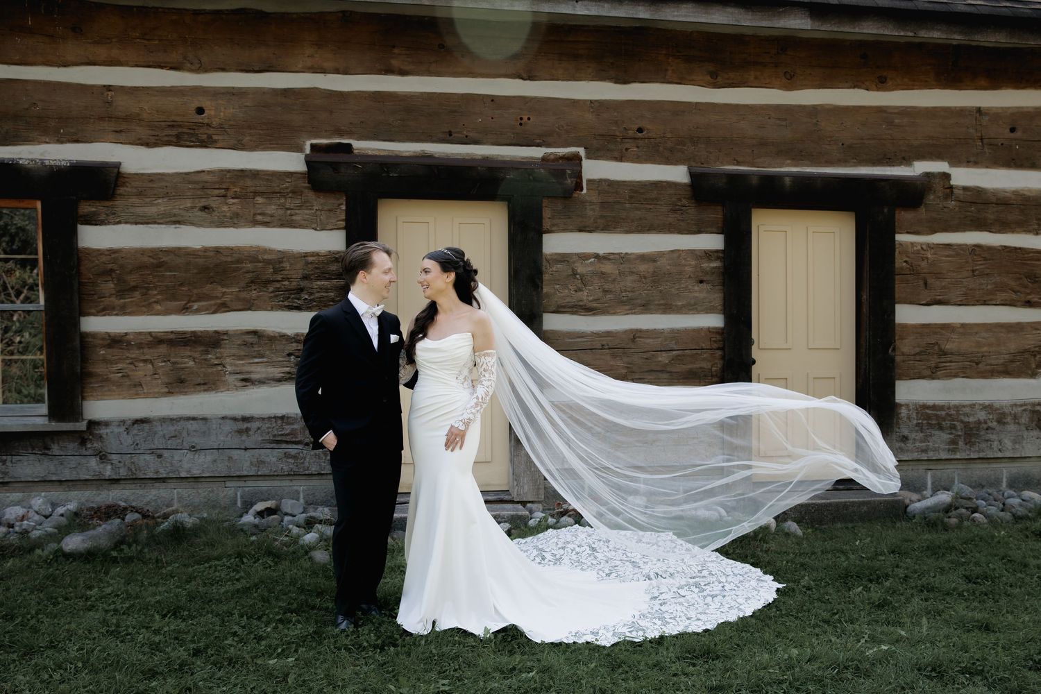 A bride in a flowing white wedding dress with a long veil walks along a rustic log cabin wall.