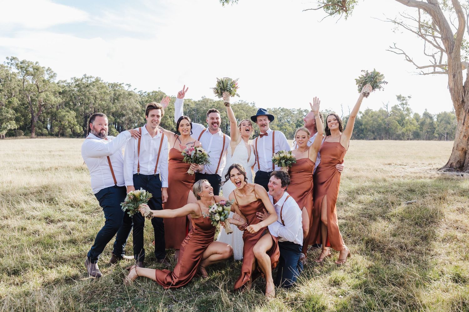 Wedding party wearing rust orange and white outfits poses together playfully in a grassy outdoor setting at sunset.