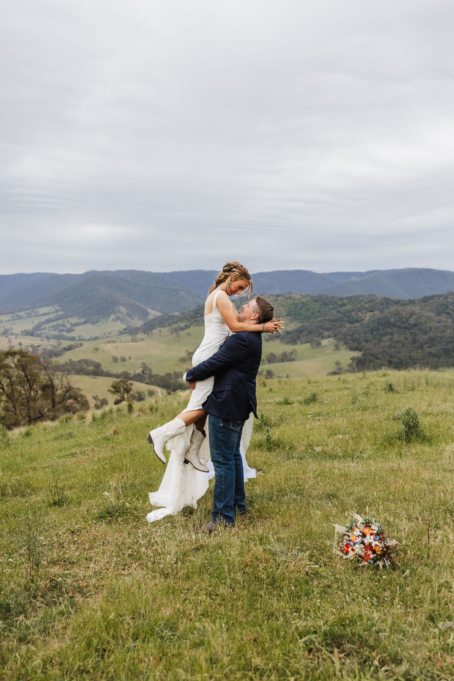 A romantic embrace amidst rolling green hills and mountains, with a colorful floral bouquet resting in the grass.