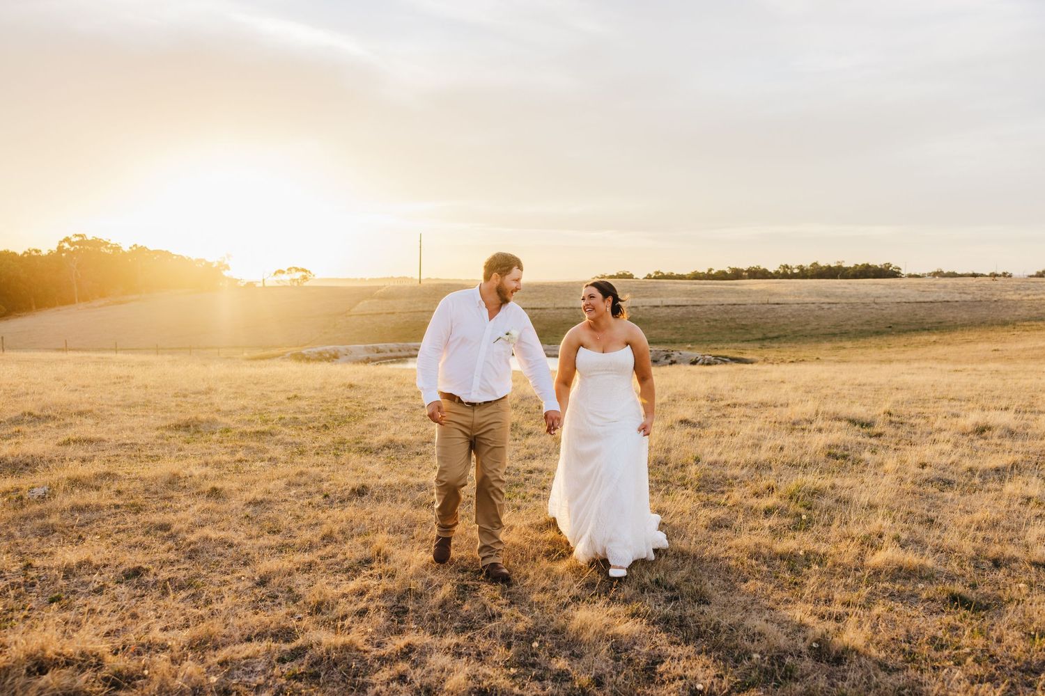 A couple holds hands while walking through a sun-drenched field at golden hour.