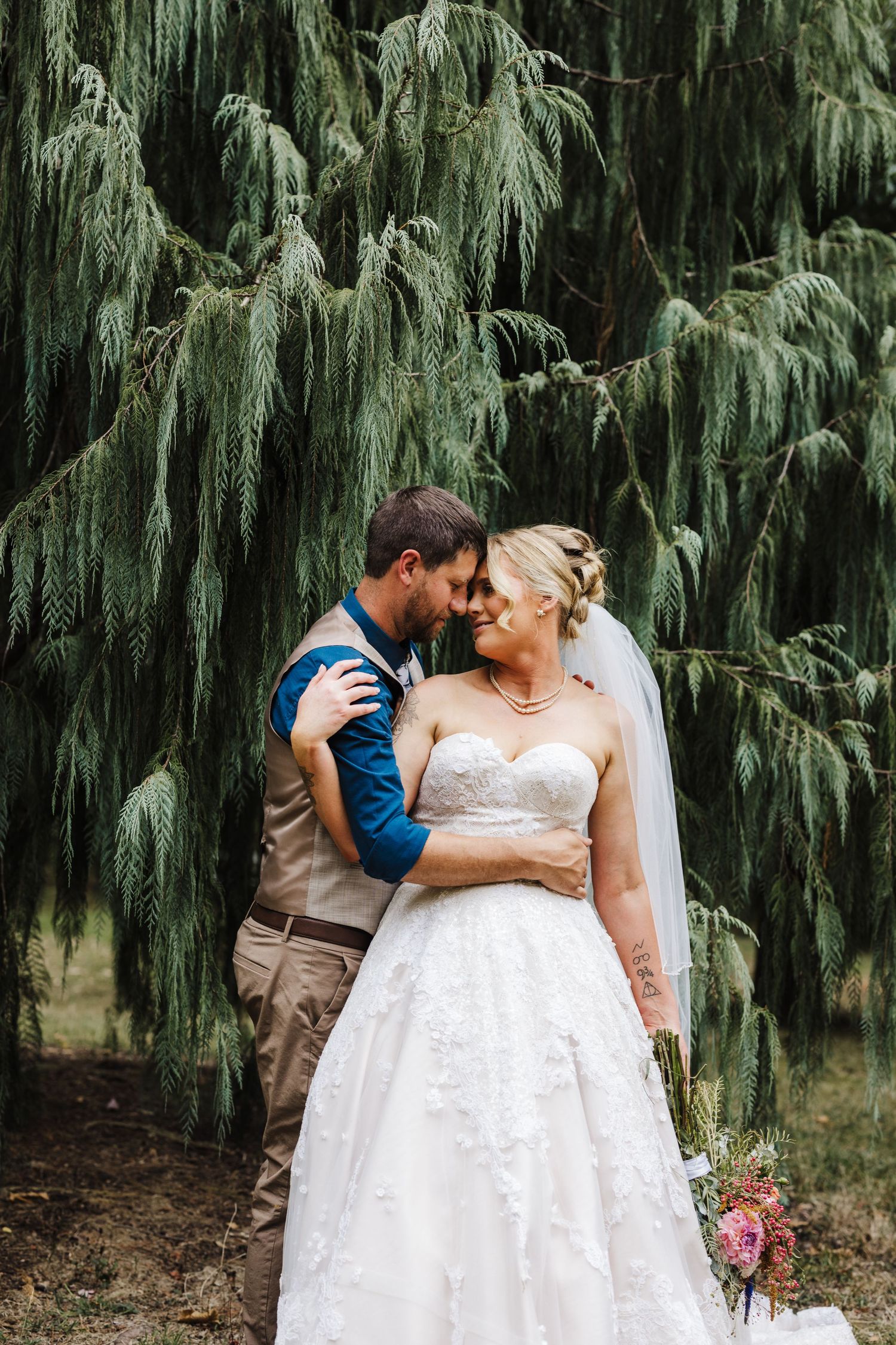 A romantic wedding couple embraces beneath a majestic weeping willow tree with trailing branches.