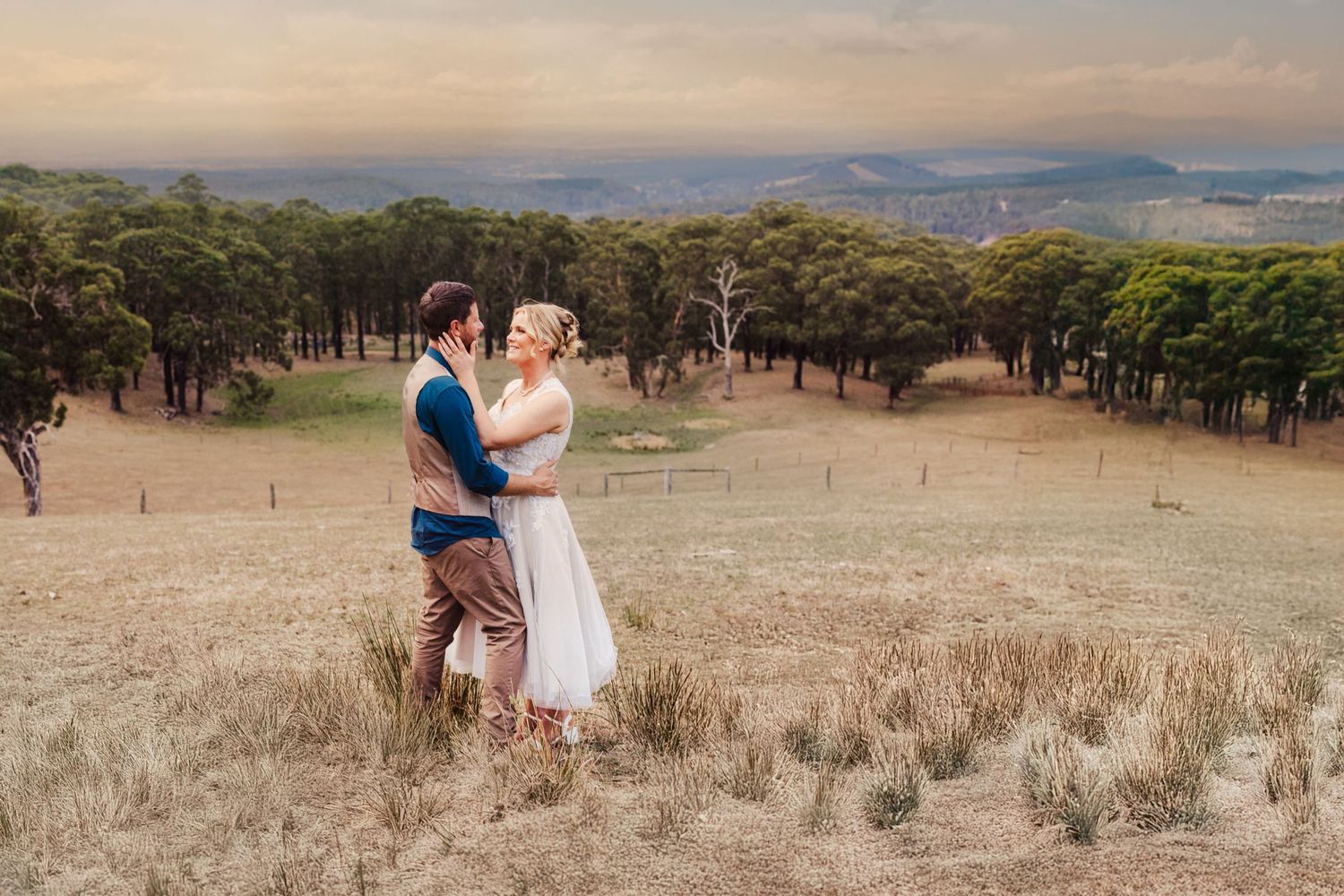 A couple embraces in their casual attire while standing in a golden grassy field with wooded hills in the background.